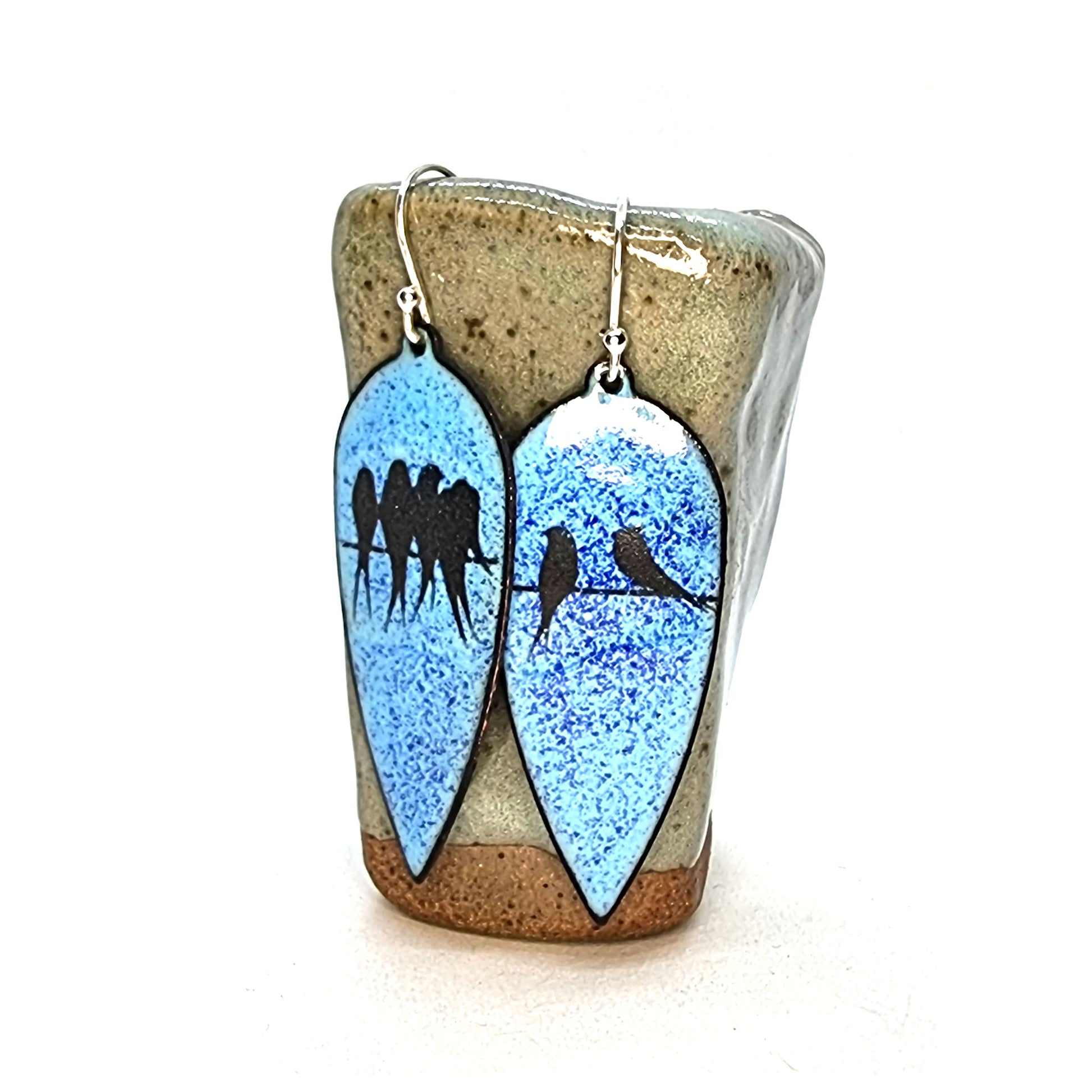 Large teardrop-shaped enamel drop earrings with black silhouettes of birds sitting on a wire against a blue background - on a vase