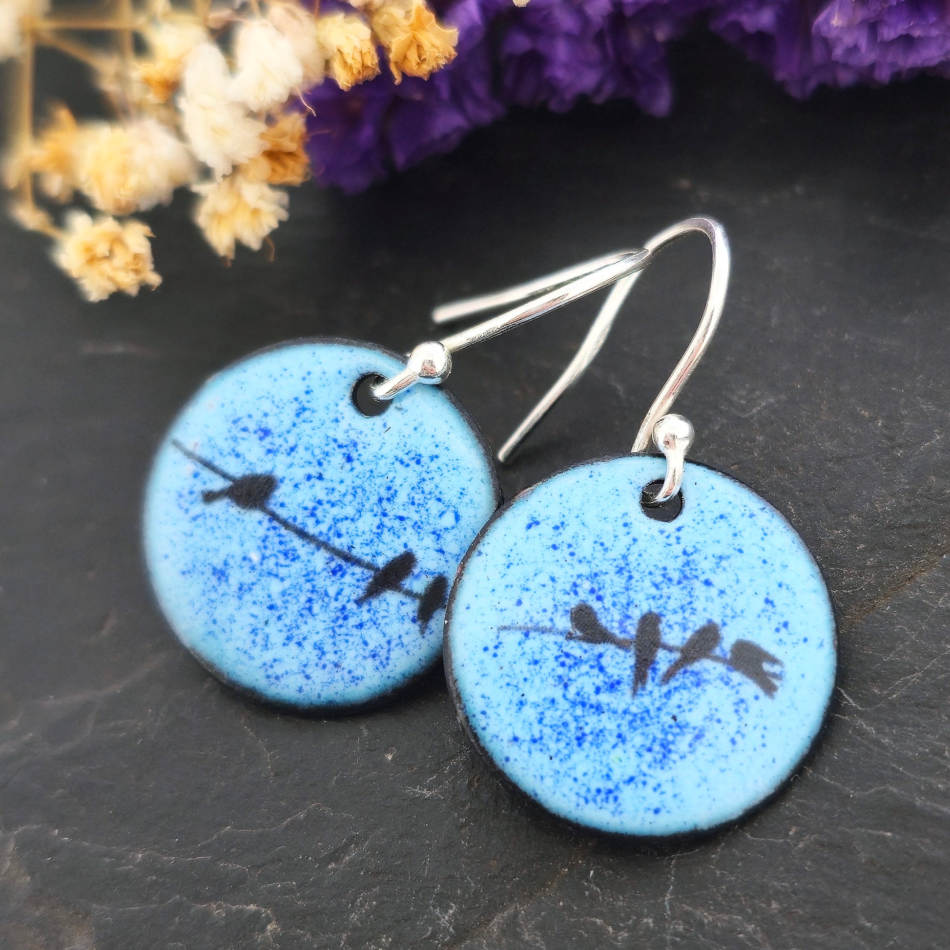 Round enamel drop earrings with blue background and black silhouettes of birds sitting on a wire. On silver ear hooks. Shown on a black background with flowers. Handmade. Mijoux Jewellery.