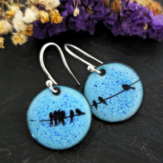 Round enamel drop earrings with blue background and black silhouettes of birds sitting on a wire. On silver ear hooks. Shown on a black background with flowers. Handmade. Mijoux Jewellery.