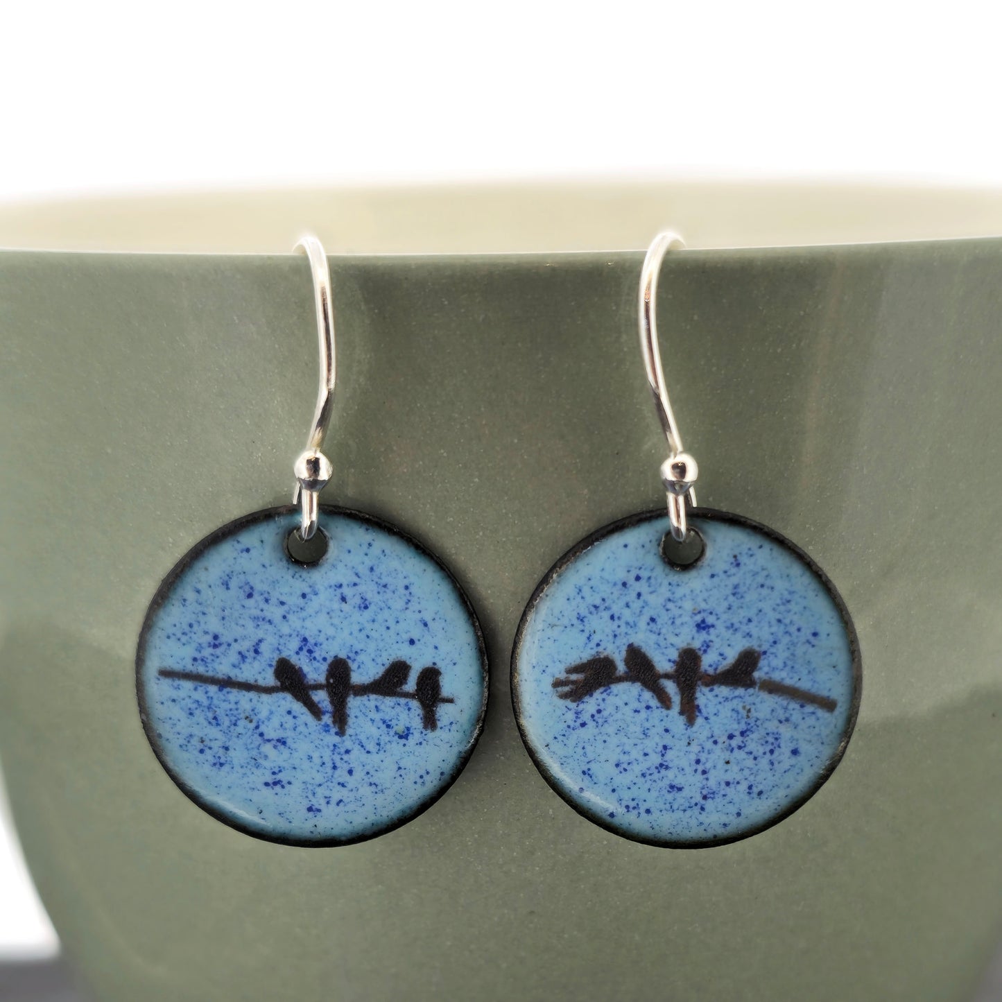 A pair of round light blue speckled drop earrings with a black bird design and silver fishhook wires, displayed against a muted green ceramic background. Handmade. Mijoux Jewellery.