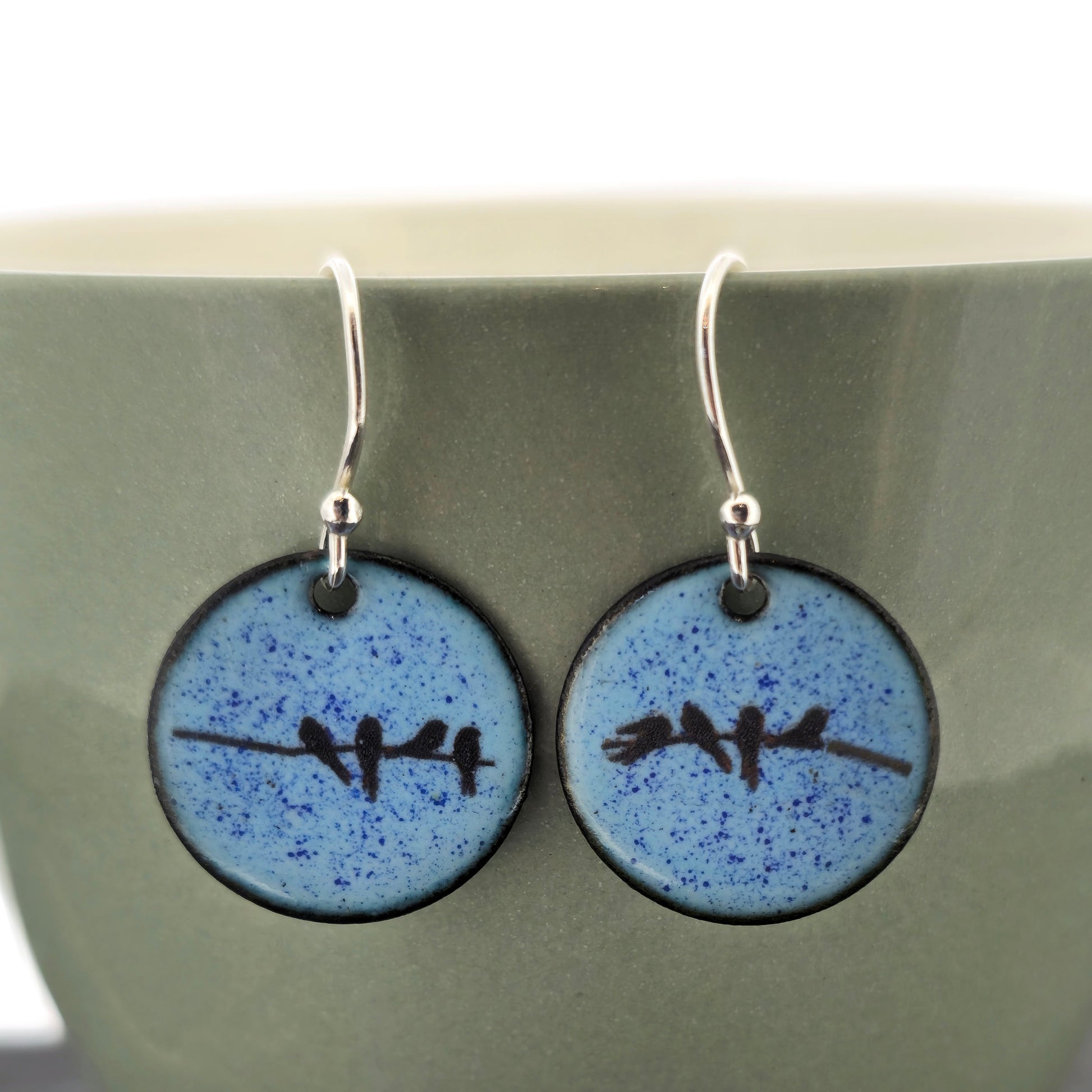 A pair of round light blue speckled drop earrings with a black bird design and silver fishhook wires, displayed against a muted green ceramic background. Handmade. Mijoux Jewellery.