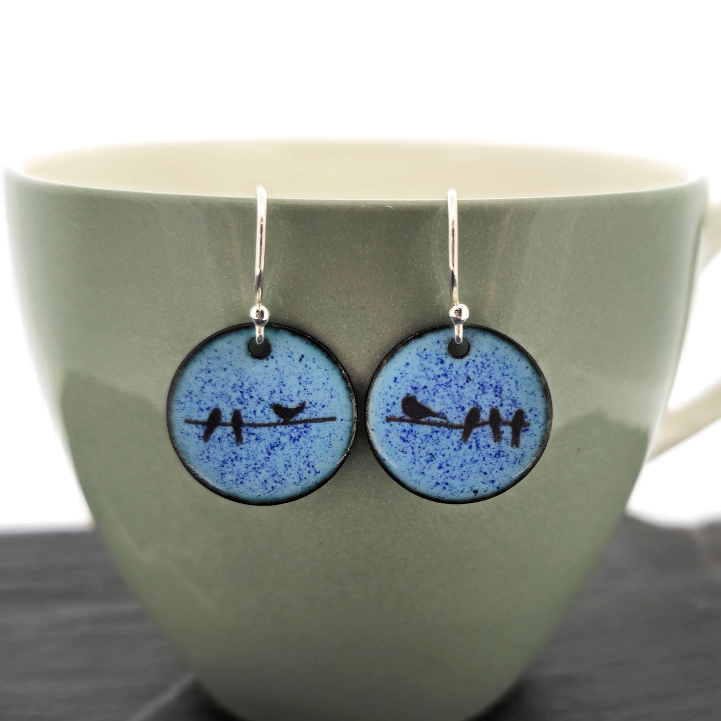A pair of round light blue speckled drop earrings with a black bird design and silver fishhook wires, displayed against a muted green ceramic background. Handmade. Mijoux Jewellery.