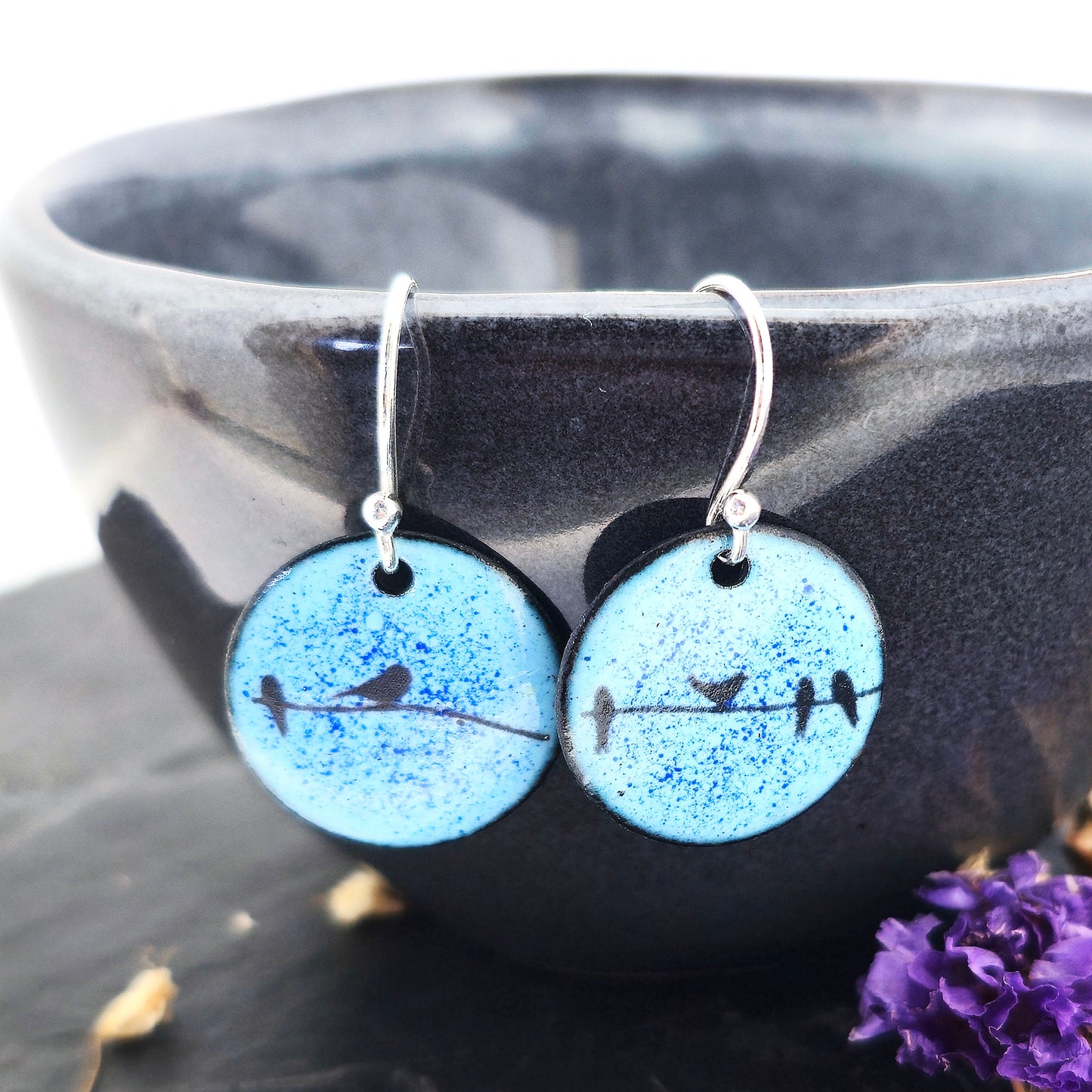 Round enamel drop earrings with blue background and black silhouettes of birds sitting on a wire. On silver ear hooks. Shown on a black bowl with flowers. Handmade. Mijoux Jewellery.