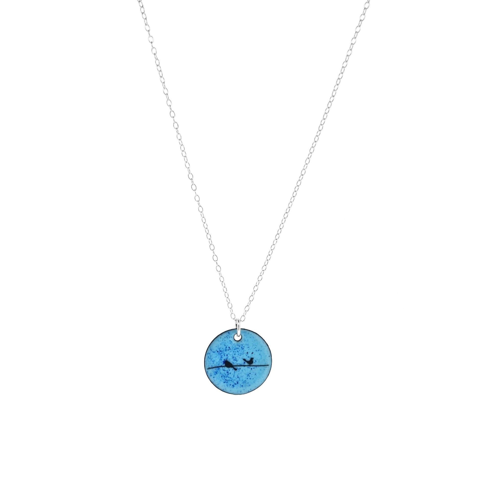 Round enamel pendant with bird silhouettes sat on a wire against a blue background. Suspended on a silver chain.