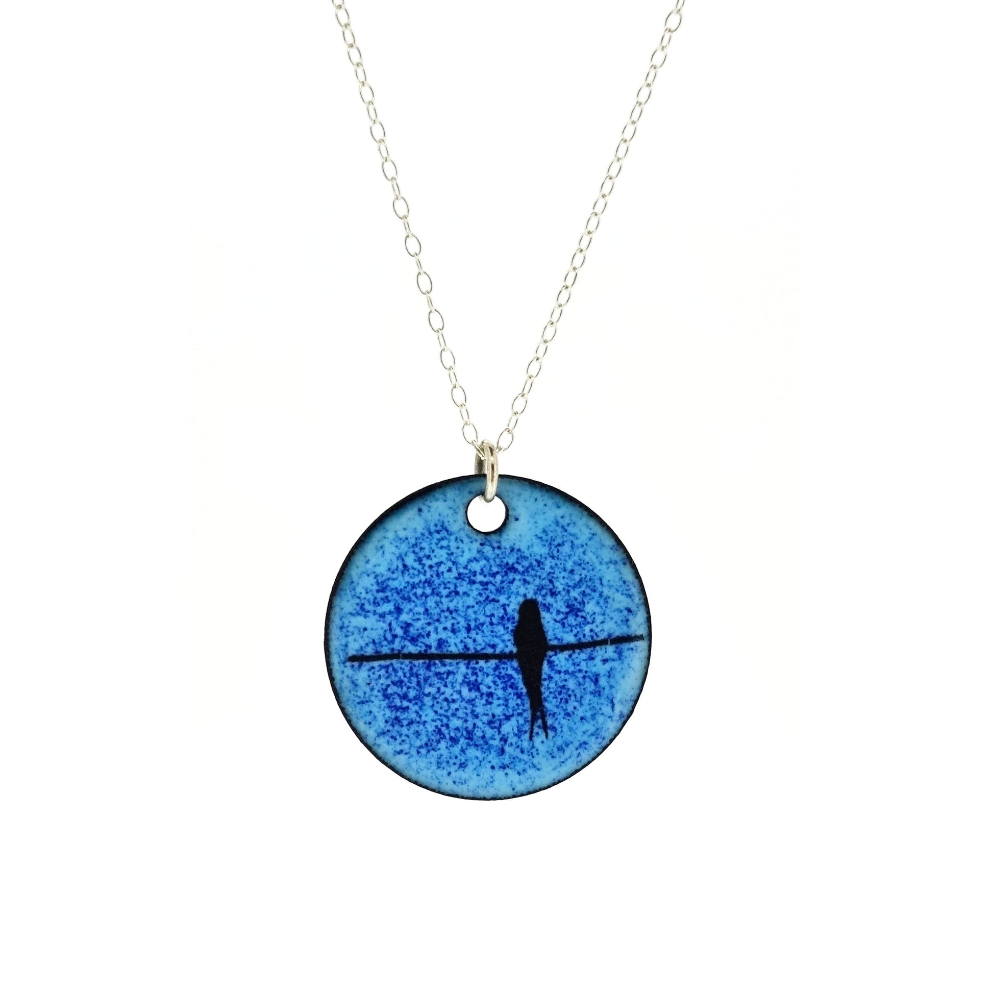 Round enamel pendant with bird silhouette sat on a wire against a blue background. Suspended on a silver chain.