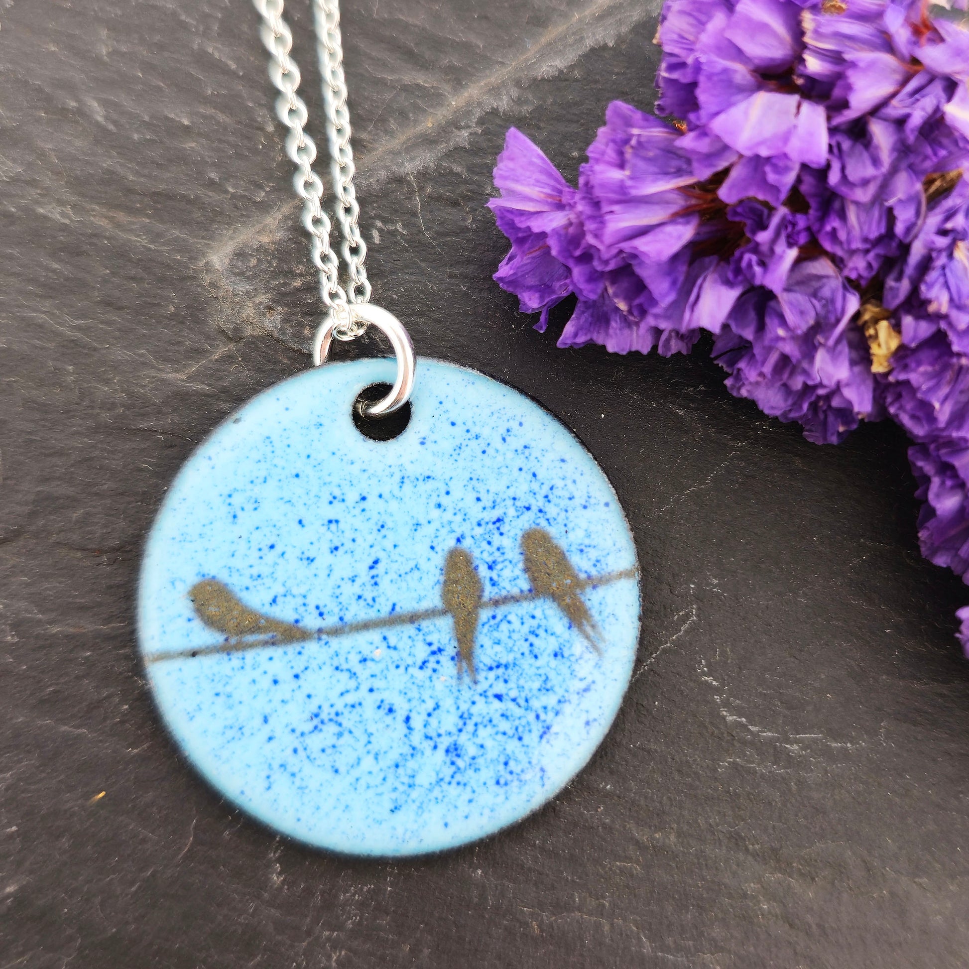 Necklace with a blue enamel pendant featuring silhouettes of birds on a wire. Shown on a dark surface with dried flowers. Mijoux Jewellery.