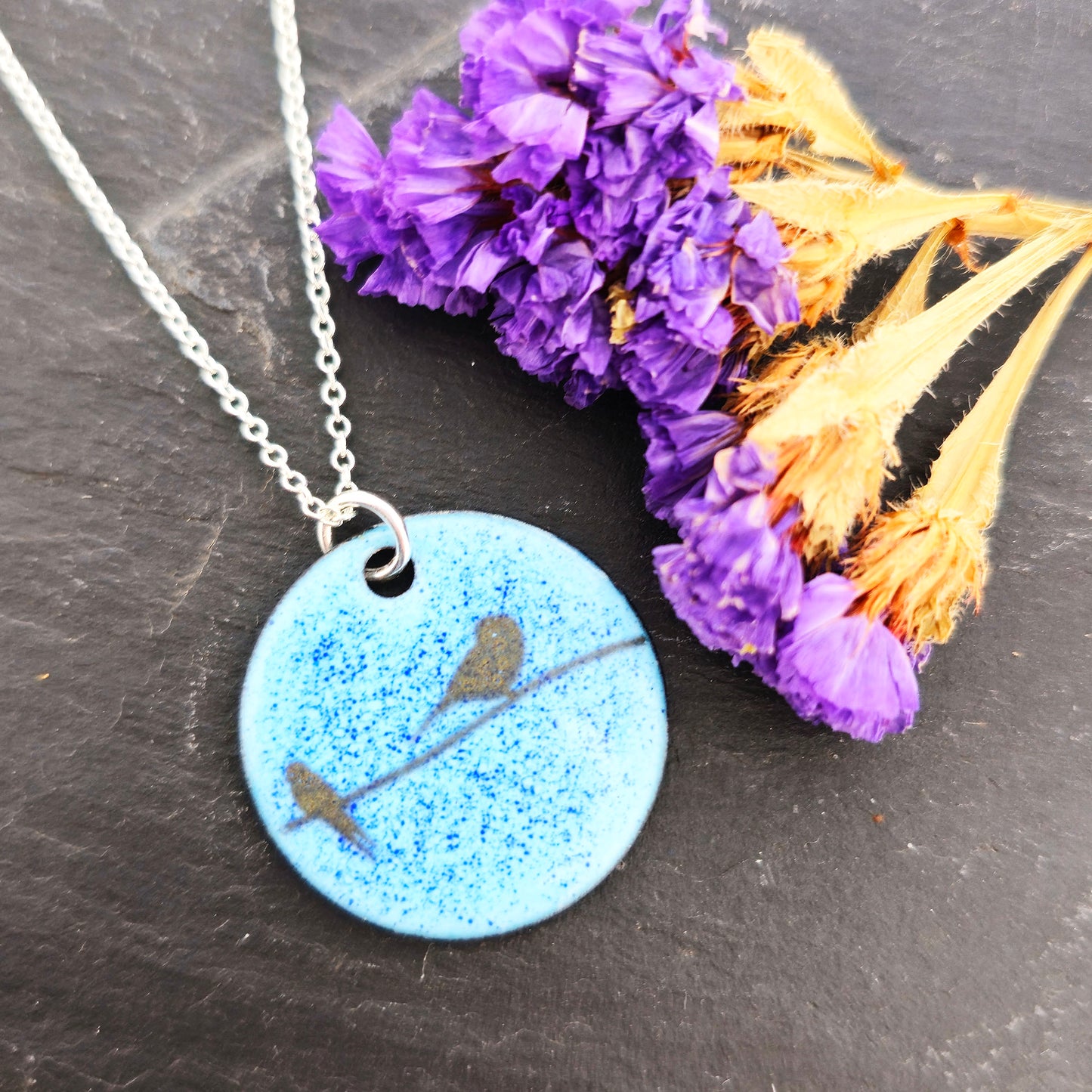 Necklace with a blue enamel pendant featuring silhouettes of birds on a wire. Shown on a dark surface with dried flowers. Mijoux Jewellery.