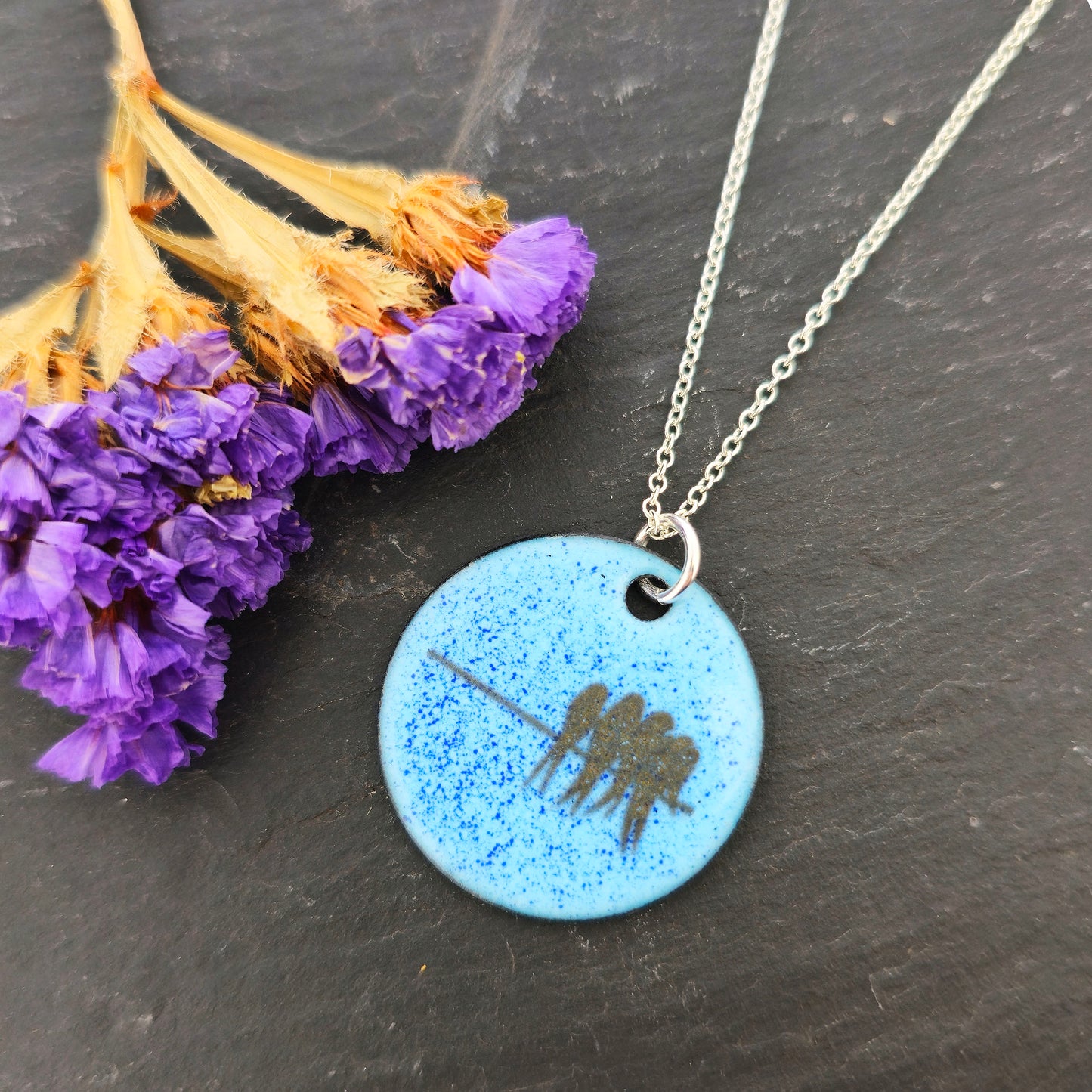 Necklace with a blue enamel pendant featuring silhouettes of birds on a wire. Shown on a dark surface with dried flowers. Mijoux Jewellery.
