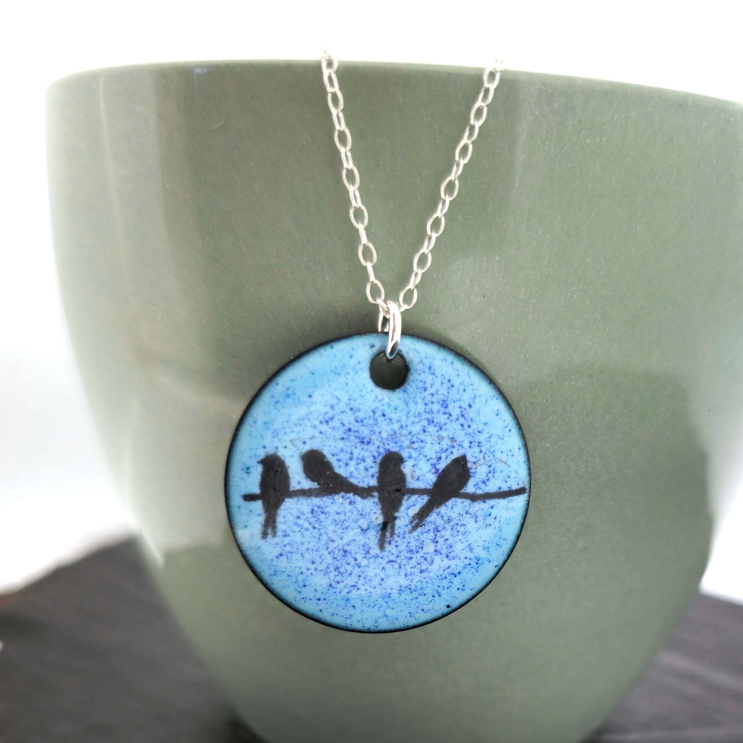 Necklace with a blue enamel pendant featuring silhouettes of birds displayed against a green ceramic background.
