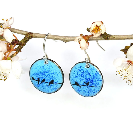 Round enamel drop earrings with blue background and black silhouettes of birds sitting on a wire. - Suspended from a blossom branch