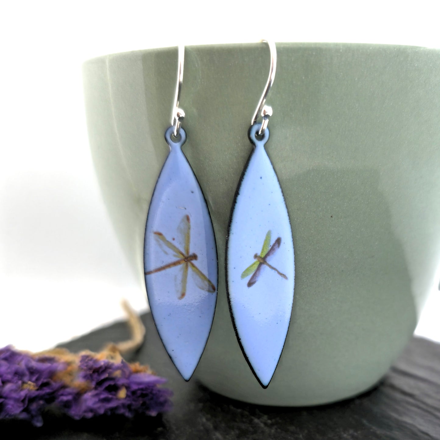 A pair of enamel earrings with different coloured dragonflies on a pale blue background. On silver ear hooks. MijouxJewellery.