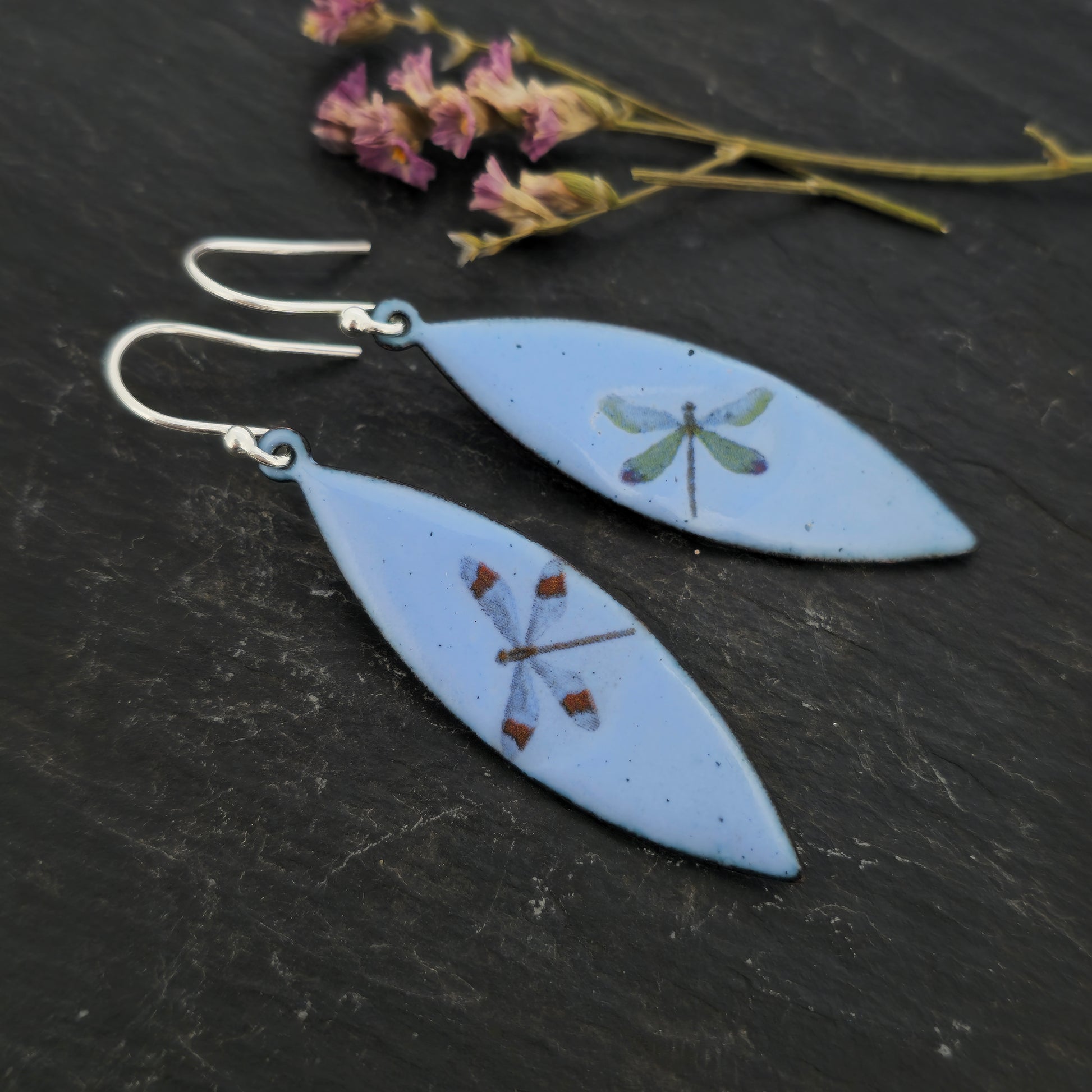 A pair of enamel earrings with different coloured dragonflies on a pale blue background. On silver ear hooks. On slate with flowers.