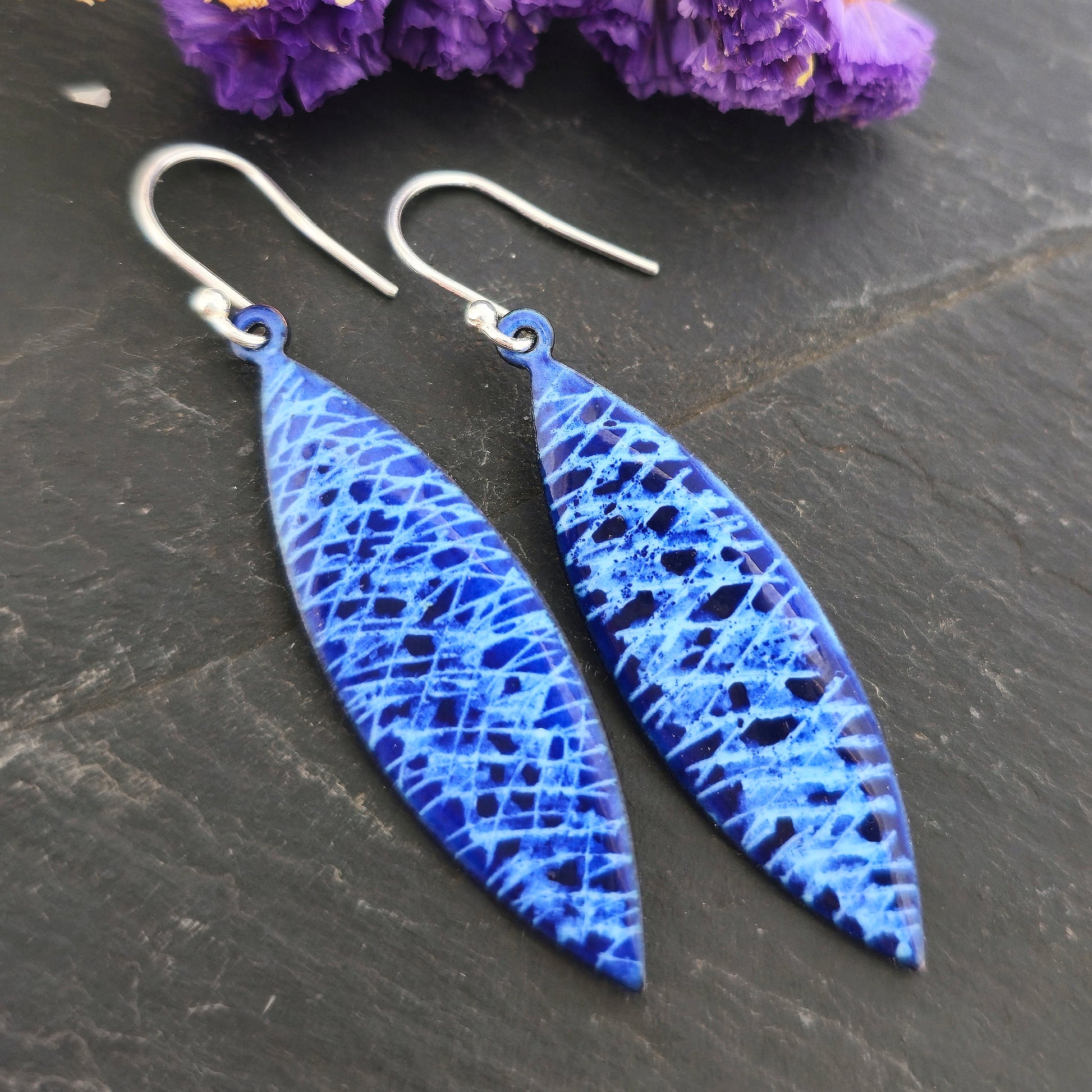 A pair of handmade enamel earrings with lines of light blue and sections of dark blue. On sterling silver ear hooks. Pictured on a black background with purple flowers. Mijoux Jewellery.