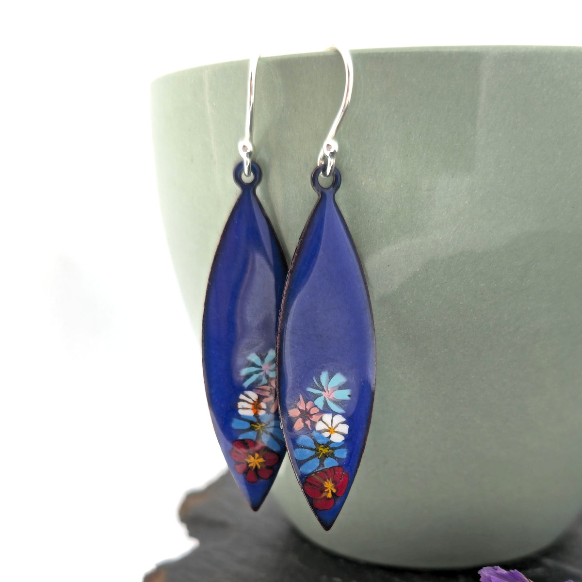 A pair of handmade boat-shaped dangling earrings with a glossy deep blue background and colorful  enamel flowers embedded near the bottom, hanging in front of a muted green ceramic cup. Mijoux jewellery.