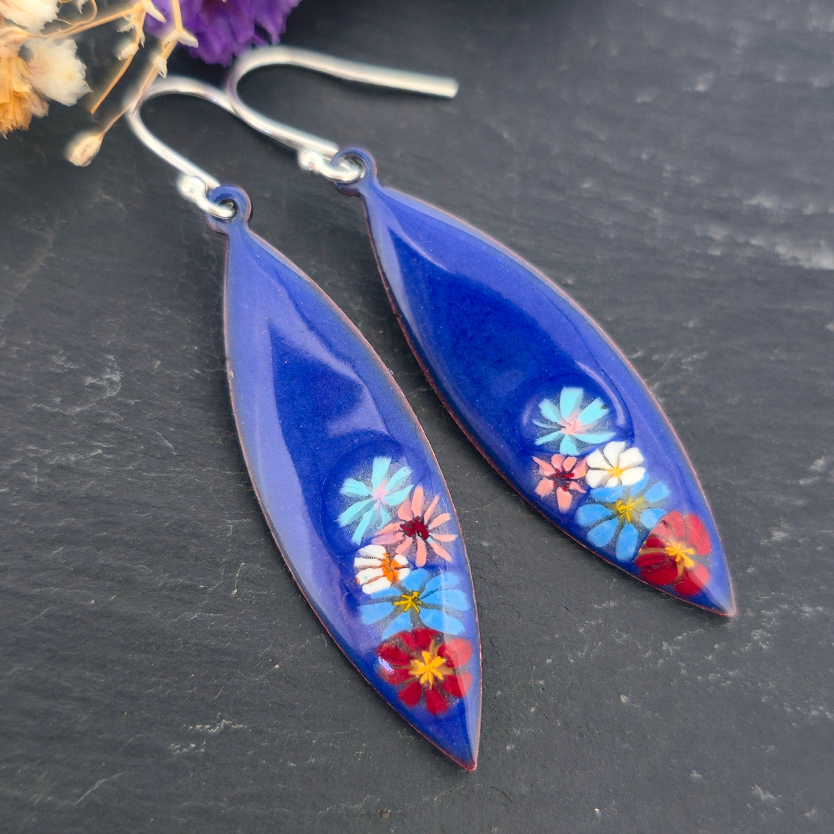 A pair of handmade boat-shaped dangling earrings with a glossy deep blue background and colorful  enamel flowers embedded near the bottom, on a black background. Mijoux jewellery.