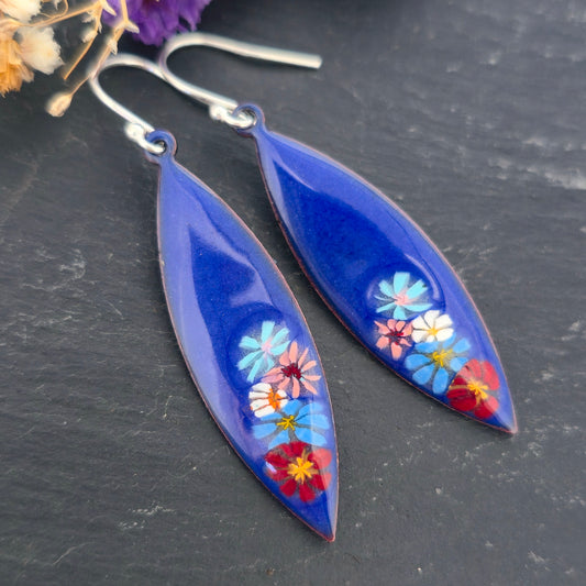 A pair of handmade boat-shaped dangling earrings with a glossy deep blue background and colorful  enamel flowers embedded near the bottom, on a black background. Mijoux jewellery.