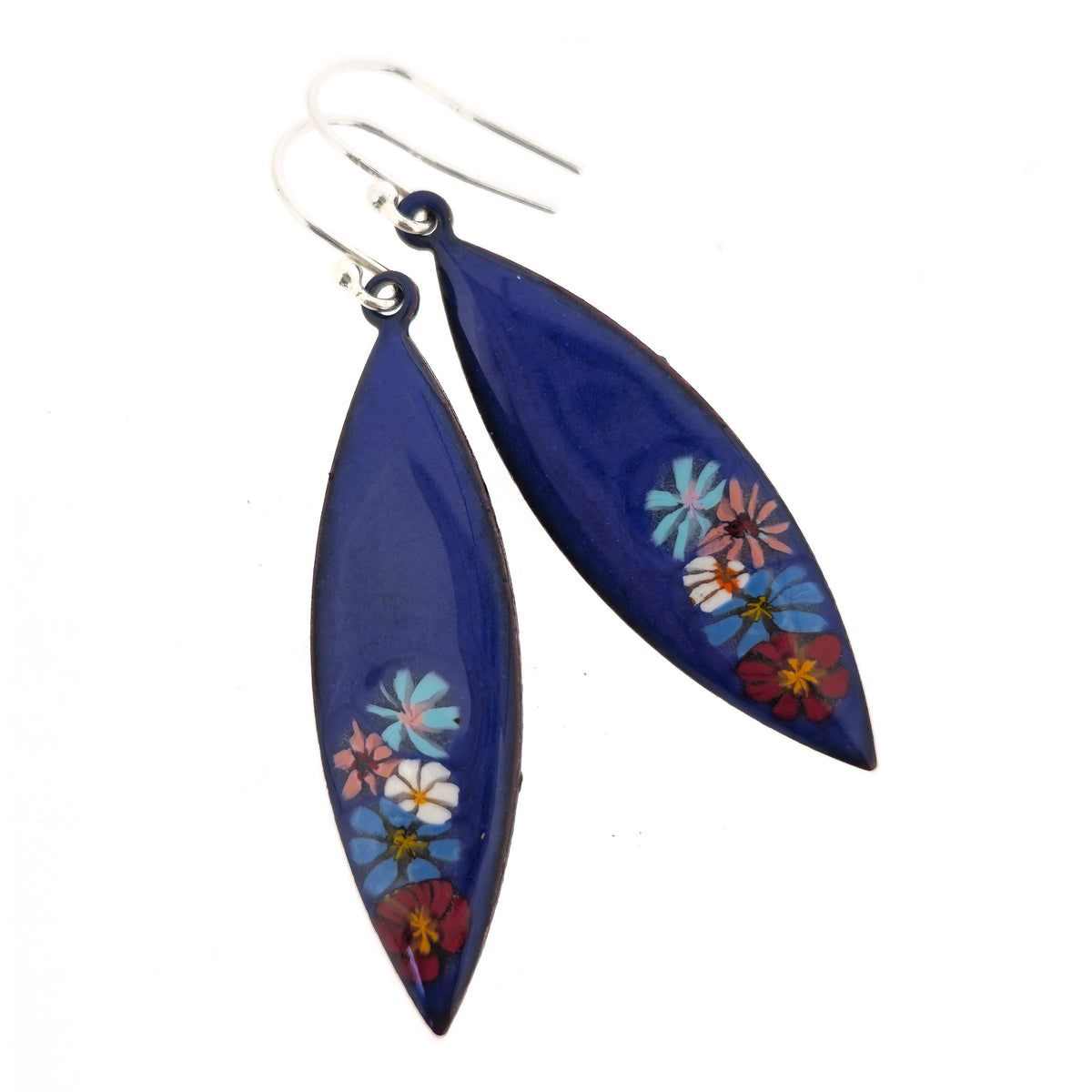 A pair of handmade blue enamel marquise shaped drop earrings on silver ear hooks. On a dark blue background are colourful enamel flowers. Shown on a white background. Mijoux jewellery.