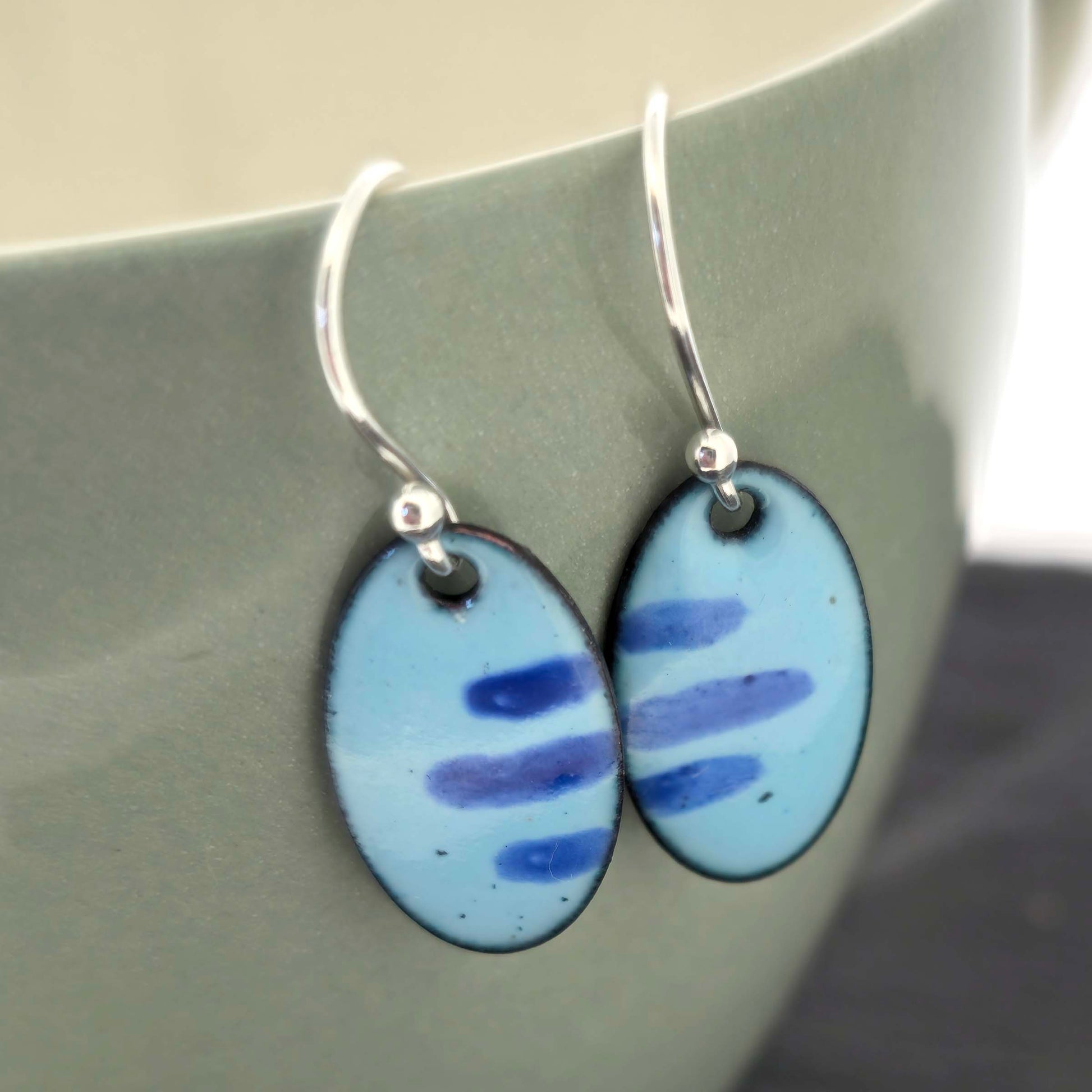 Pair of blue enamel oval earrings with a stripe pattern and silver ear hooks on a sage surface