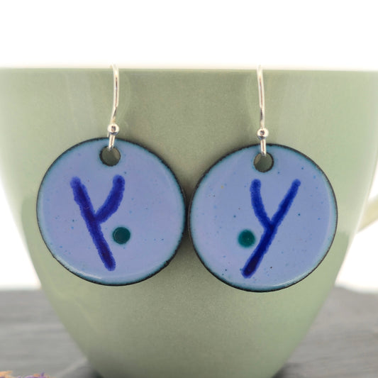 Pair of handmade enamel round drop earrings with dark blue 'y' symbols on a light blue background and green dots, hanging in front of a green mug. Mijoux jewellery.