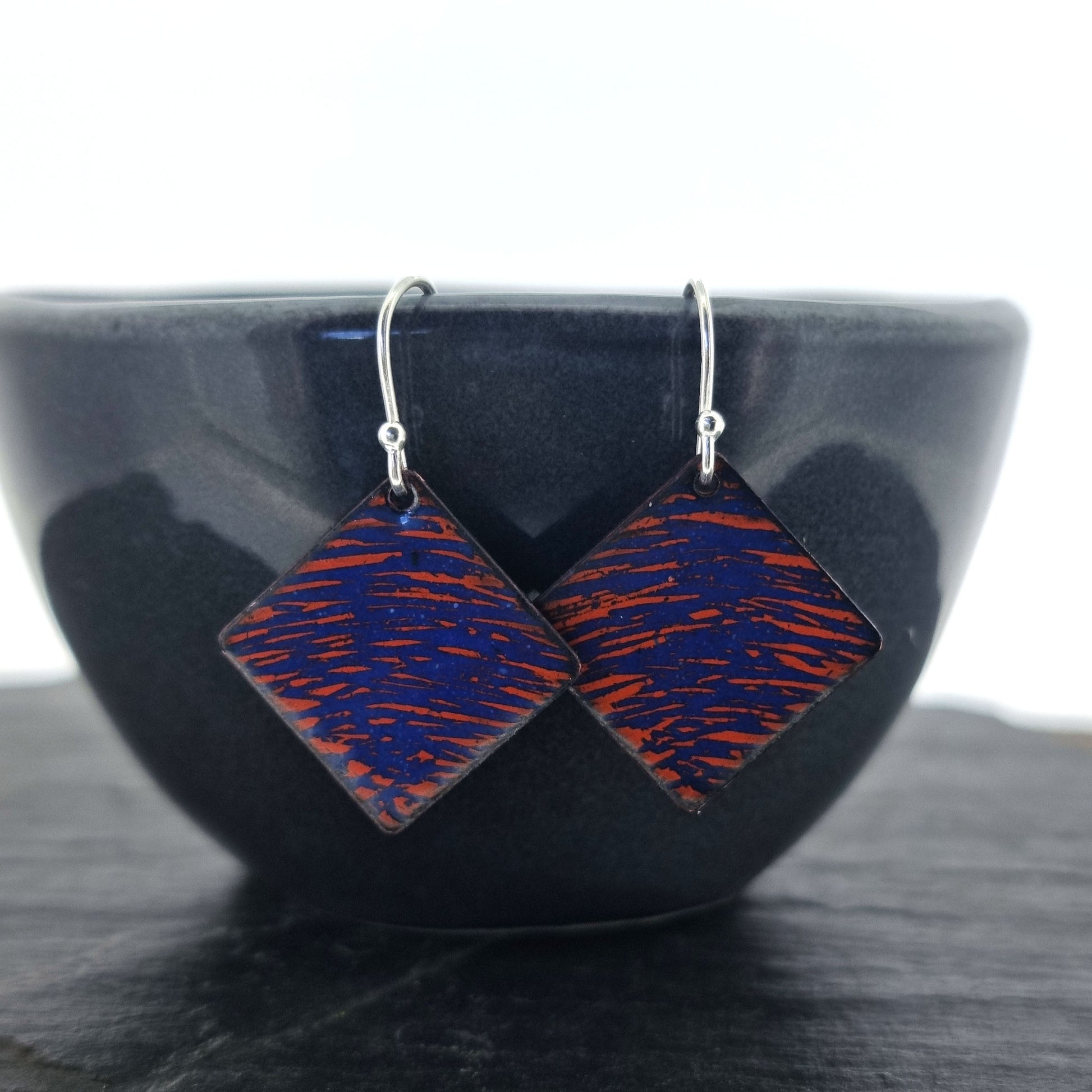 Square (diamond shaped) drop earrings with a rustic line pattern of dark blue and orange enamel. On silver ear hooks. Pictured on bowl.