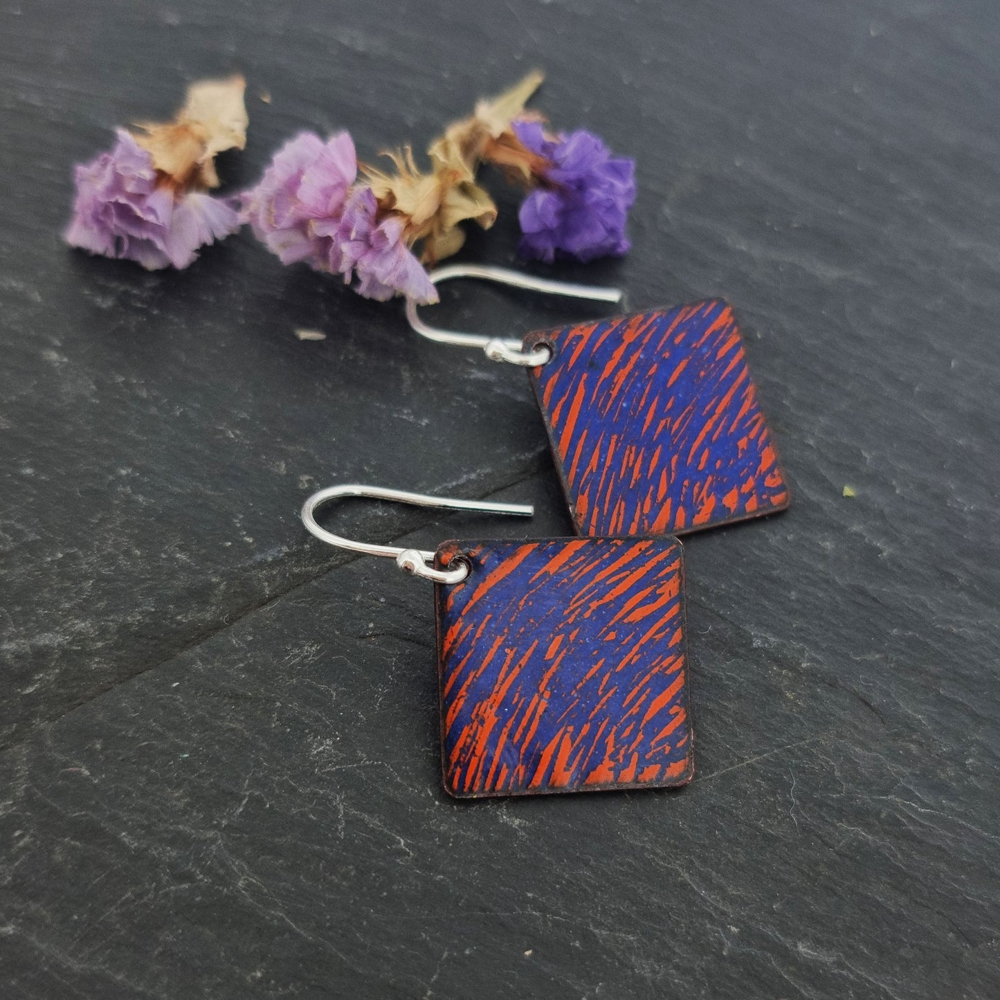 Square (diamond shaped) drop earrings with a rustic line pattern of dark blue and orange enamel. On silver ear hooks. Pictured with flowers.