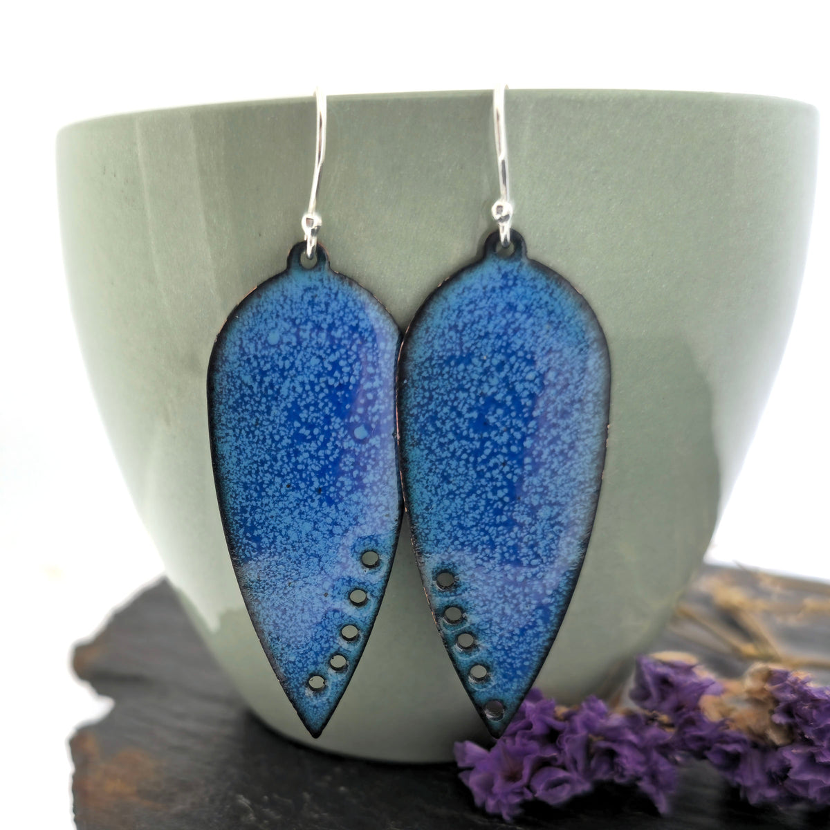 A pair of handmade teardrop-shaped blue enamel dangle earrings with silver fishhook wires and small circle cutouts along the lower edge, shown against a sage green cup. Mijoux jewellery.