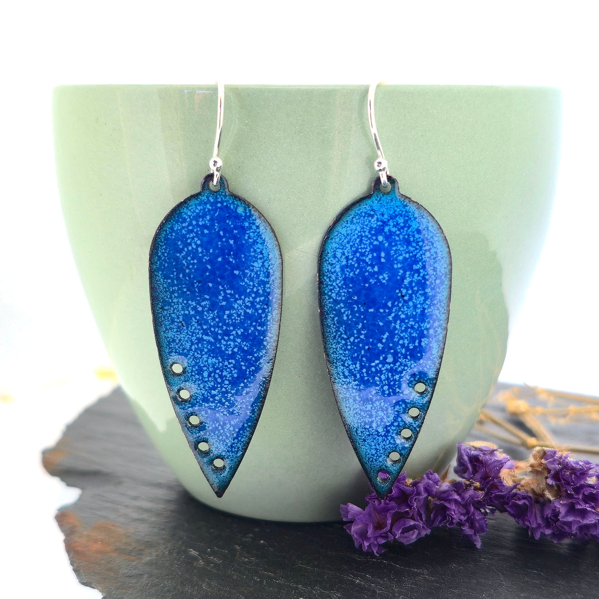 A pair of handmade teardrop-shaped blue enamel dangle earrings with silver fishhook wires and small circle cutouts along the lower edge, shown against a sage green cup. Mijoux jewellery.
