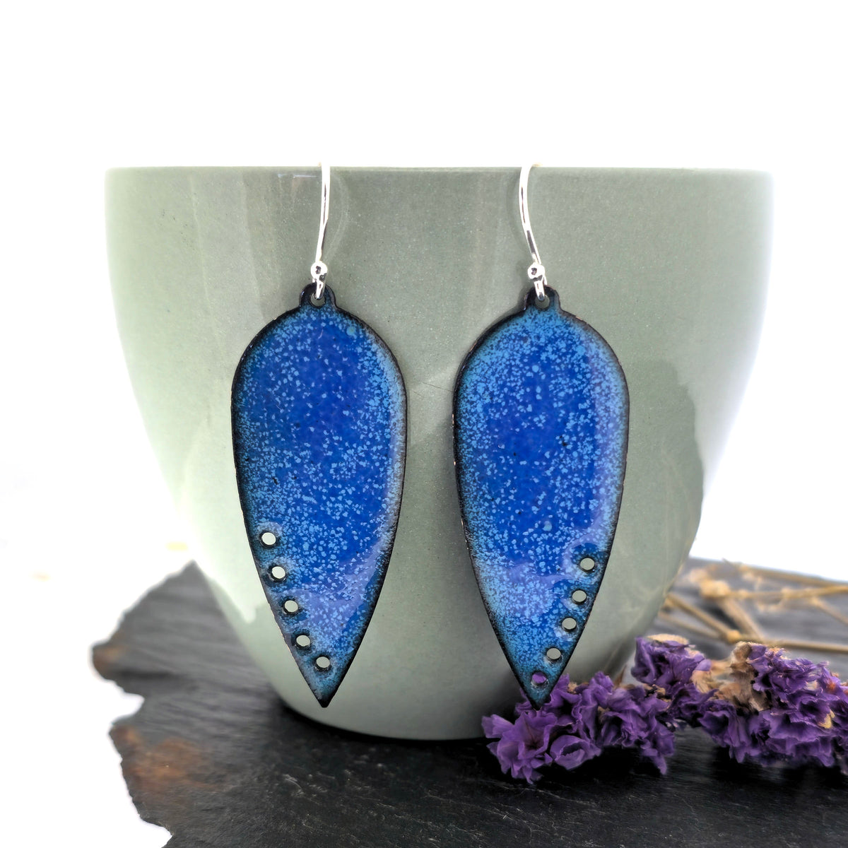 A pair of handmade teardrop-shaped blue enamel dangle earrings with silver fishhook wires and small circle cutouts along the lower edge, shown against a sage green cup. Mijoux jewellery.