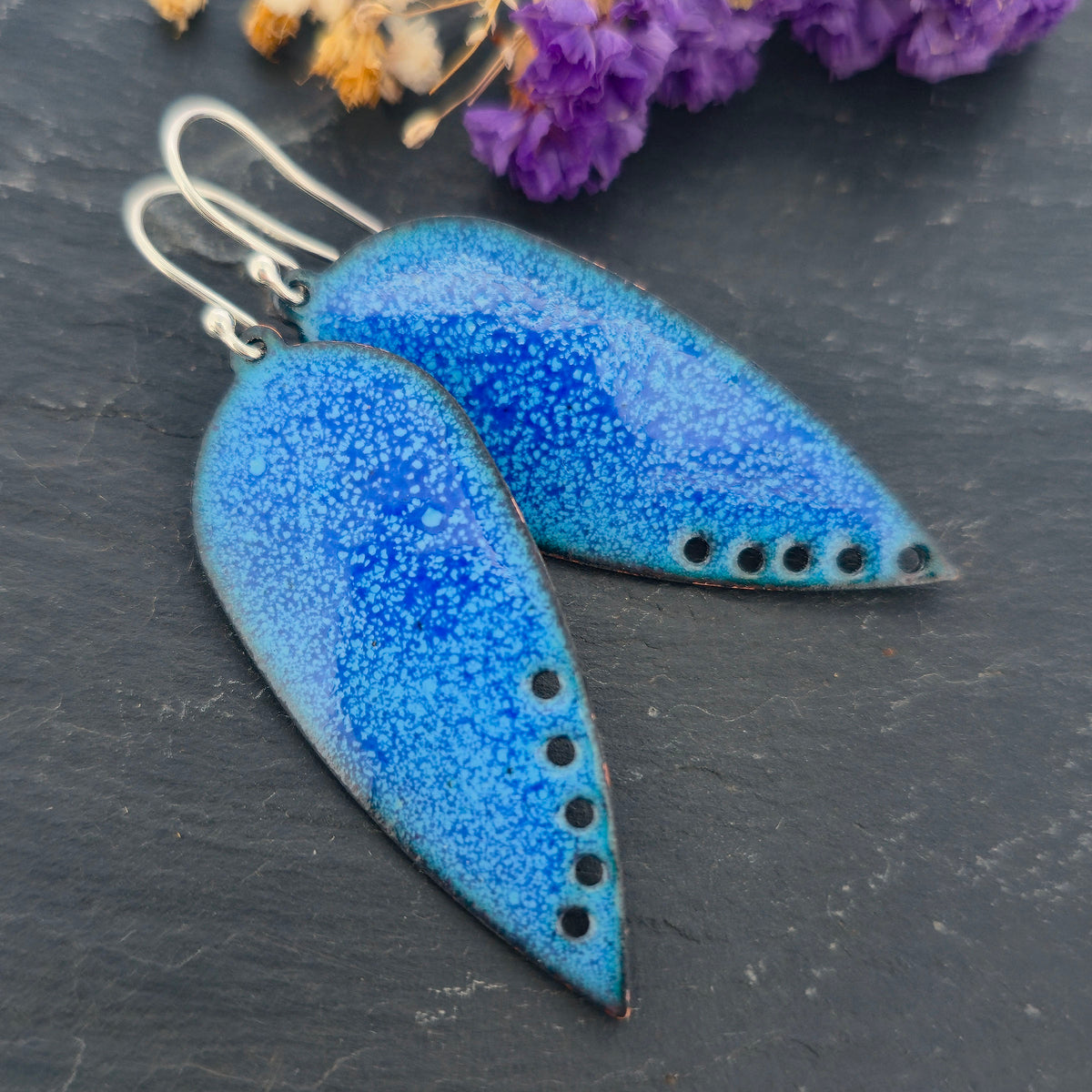 A pair of handmade teardrop-shaped blue enamel dangle earrings with silver fishhook wires and small circle cutouts along the lower edge, shown against a black background. Mijoux jewellery.