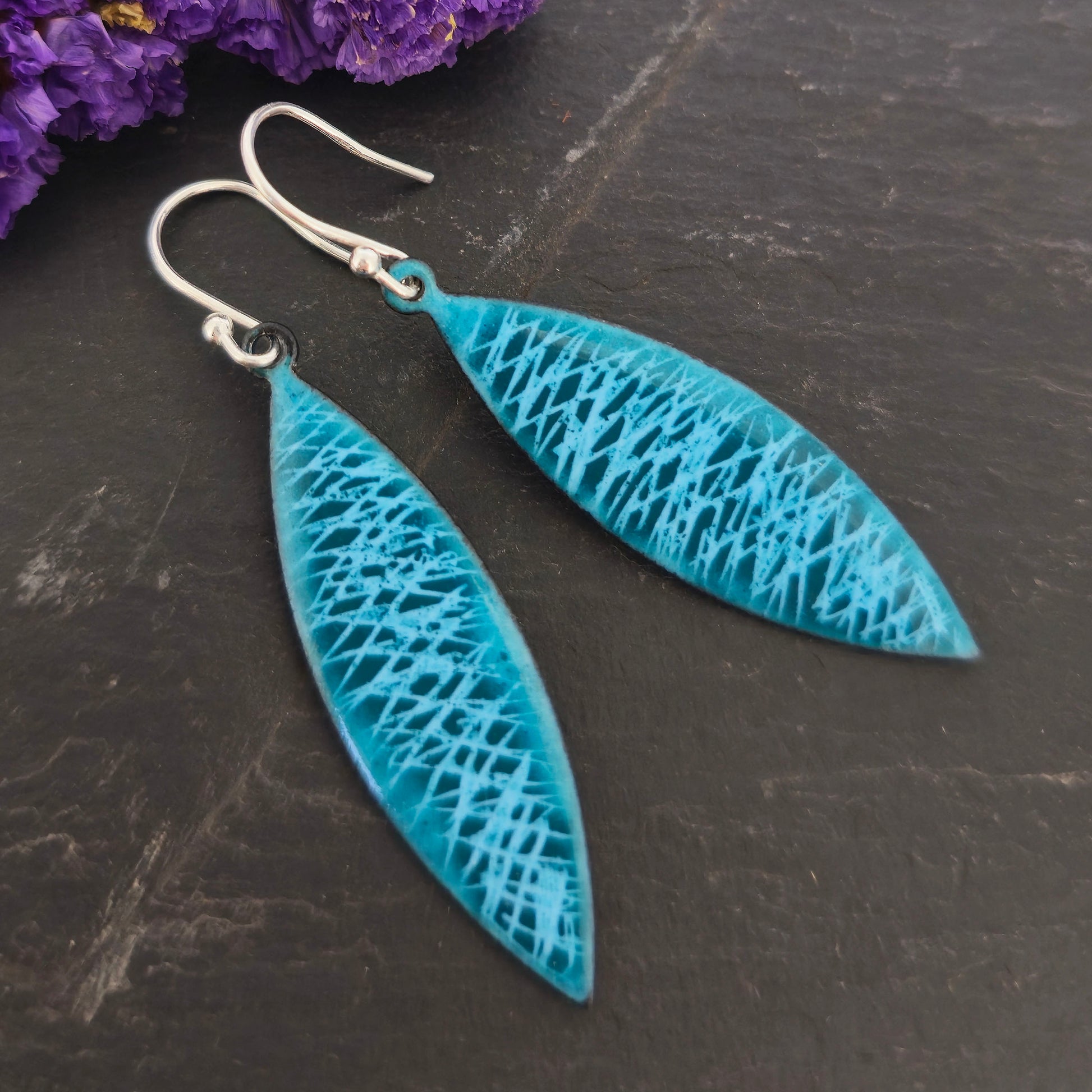 Boat-shaped drop earrings featuring a scatter line pattern in light blue and teal enamel. Suspended from silver ear hooks. Pictured on a black background with purple flowers. Mijoux jewellery.