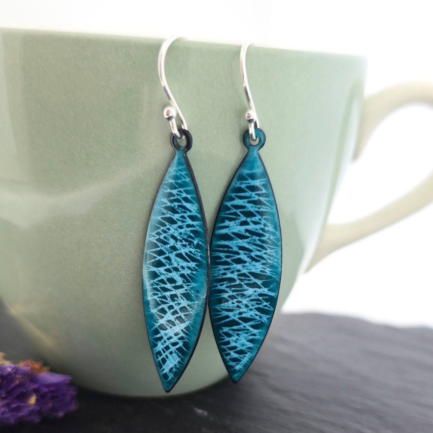 Boat-shaped drop earrings featuring a scatter line pattern in light blue and teal enamel. Suspended from silver ear hooks. Pictured on a pale green cup with purple flowers. Mijoux jewellery.