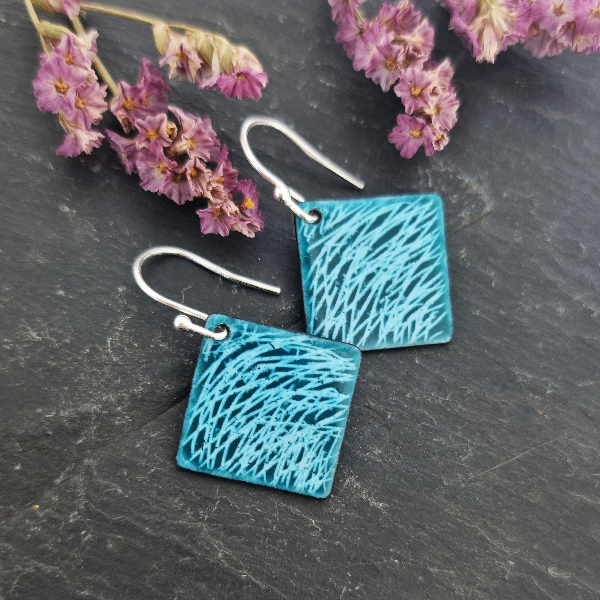 Square teal enamel earrings with lines of light blue enamel. On silver ear hooks. Pictured on slate with flowers.