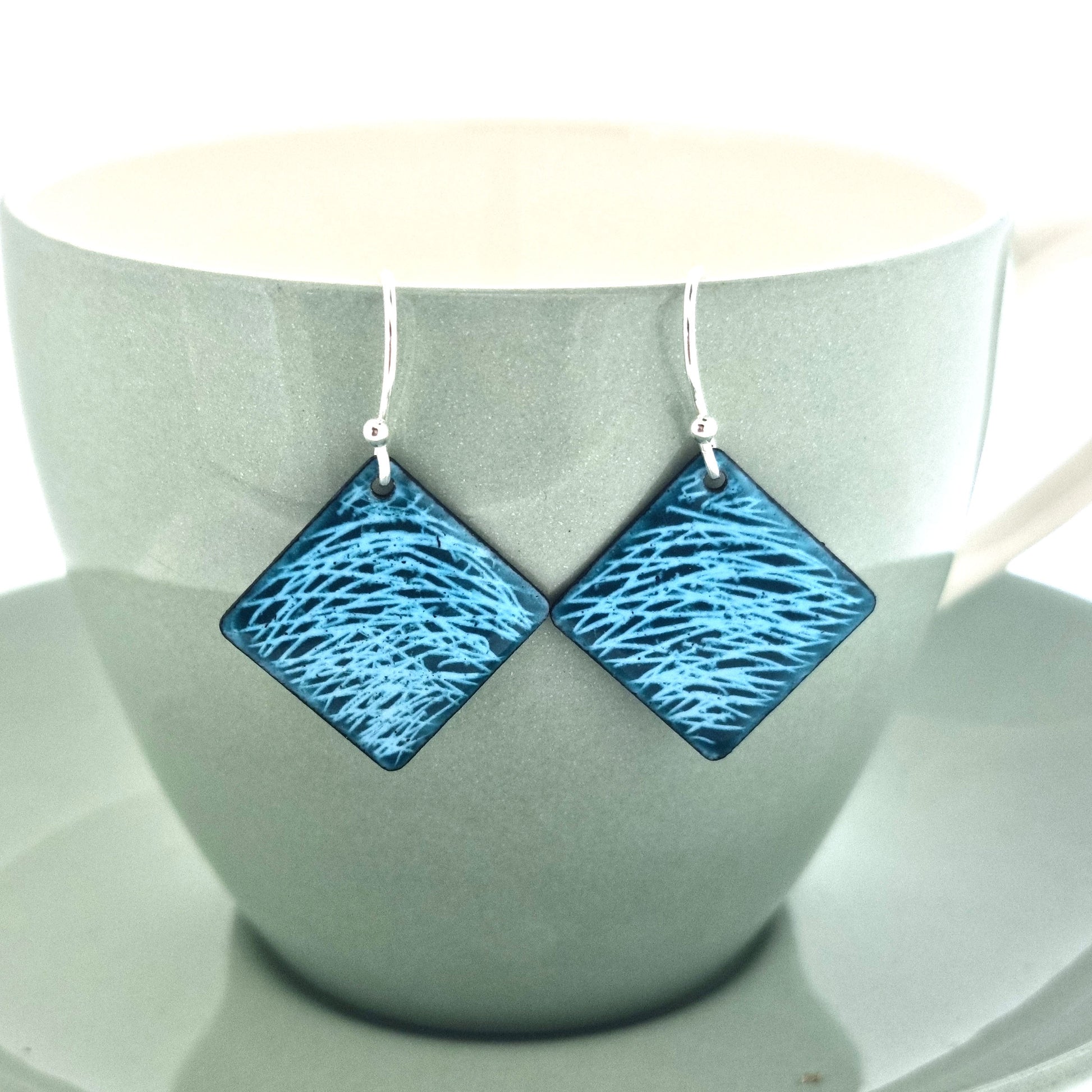 Square teal enamel earrings with lines of light blue enamel. On silver ear hooks. Pictured on a cup.