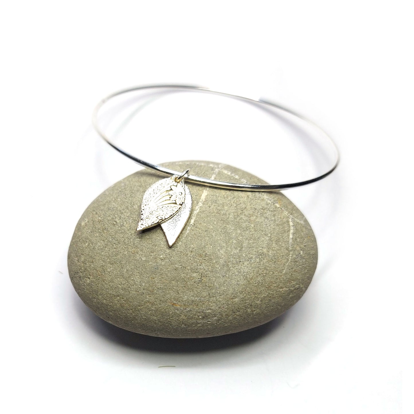 A round silver bangle with a double leaf-shaped patterned charm. On a pebble.