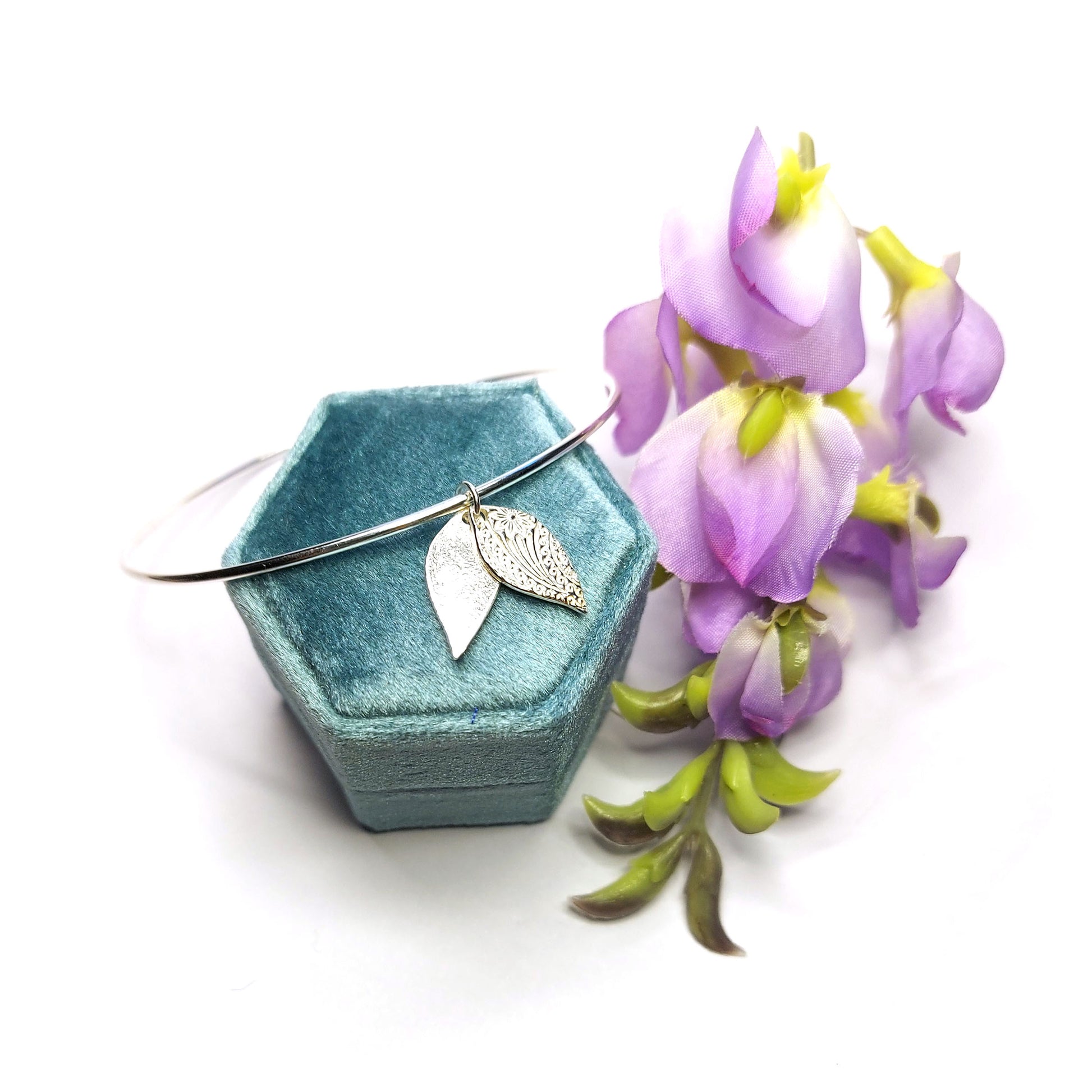 A round silver bangle with a double leaf-shaped patterned charm. On a velvet box with flowers.