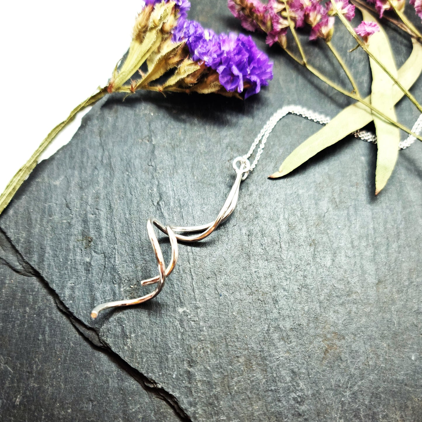 A silver twist pendant with 2 strands intertwined suspended from a silver chain. Shown on slate with flowers