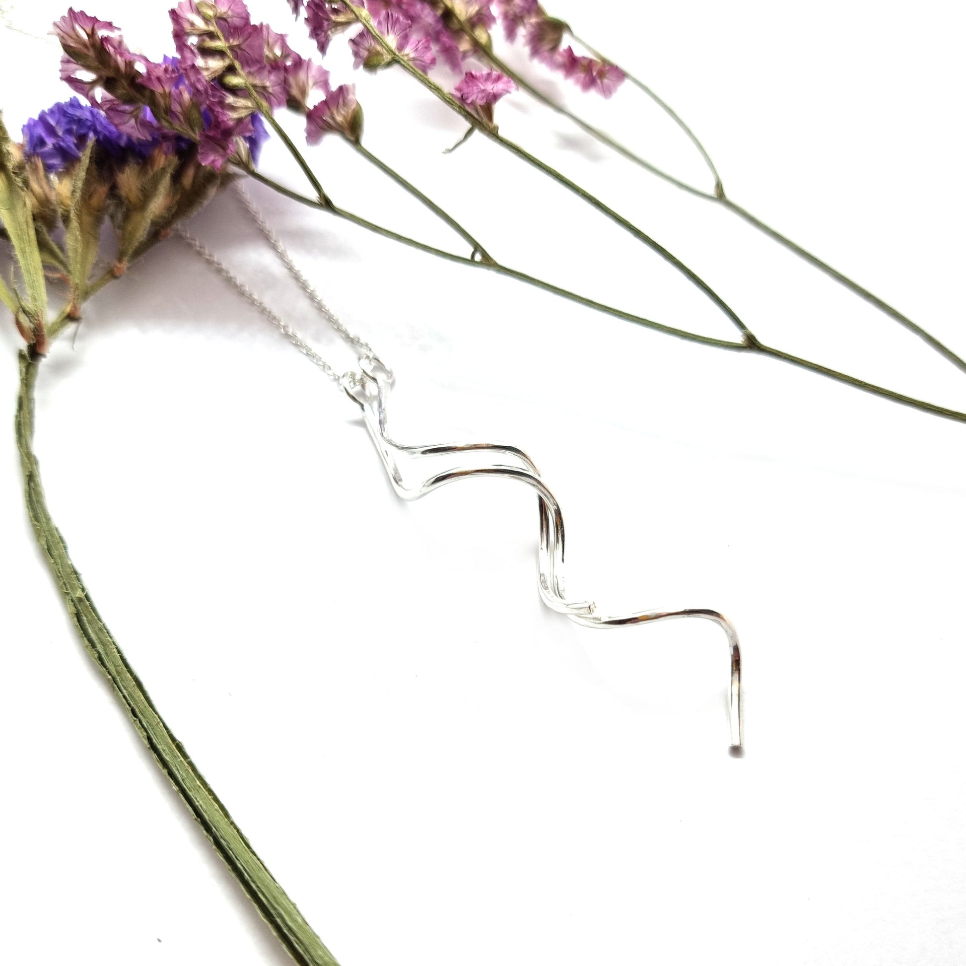 A silver spiral pendant with 2 strands intertwined suspended from a silver chain. Shown with flowers.