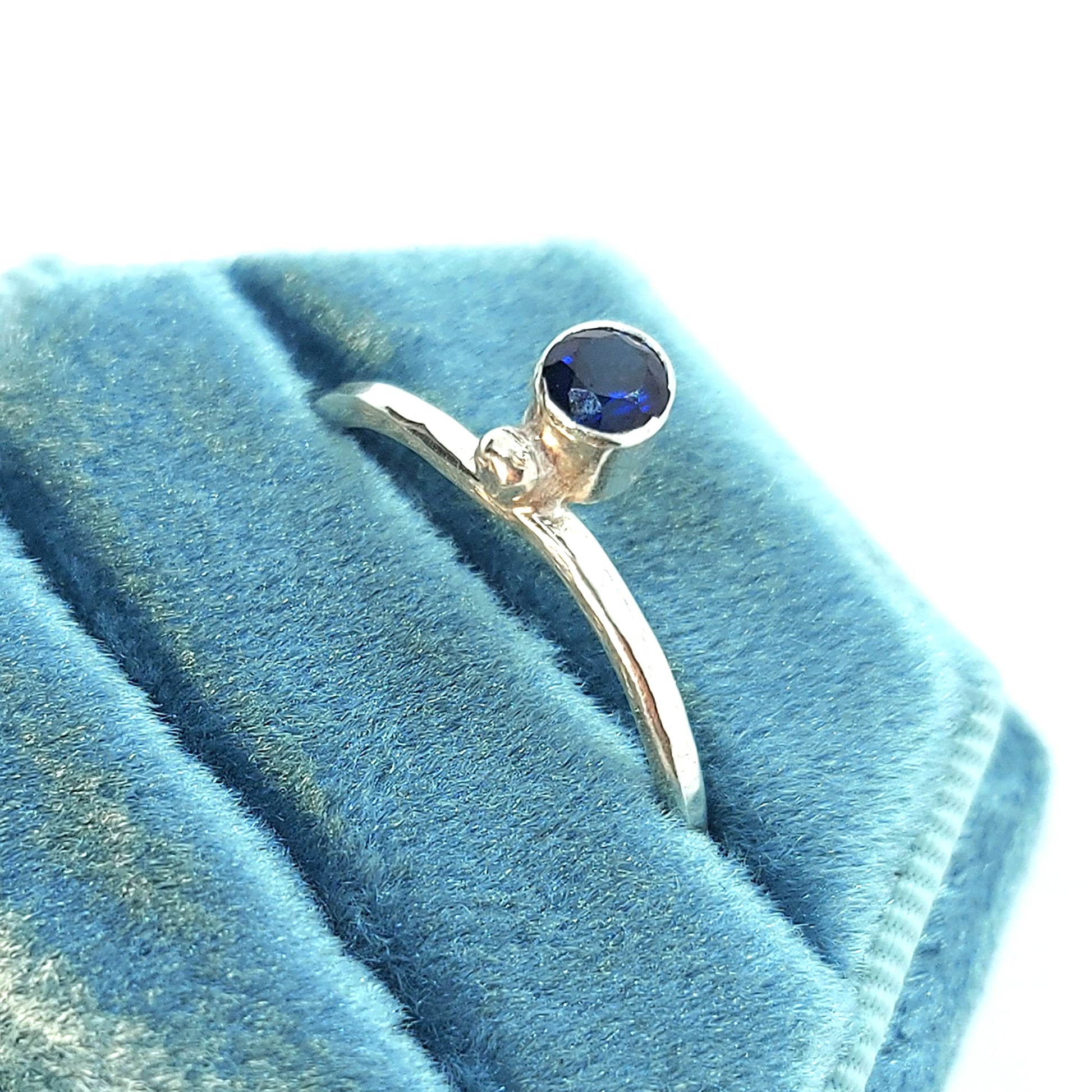 A silver thin ring with a hammered finish. The ring has a dark blue sapphire bezel set alongside a silver dot.