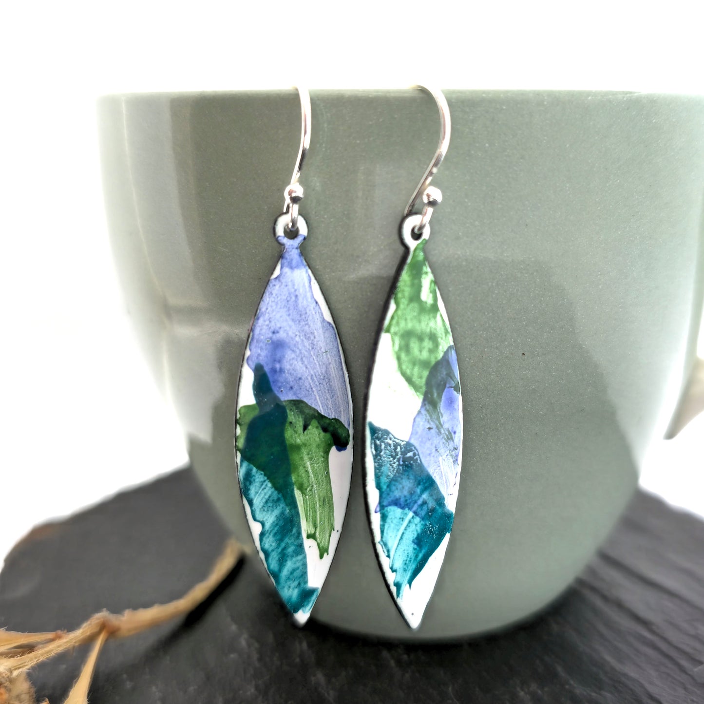 Handmade navette shaped enamel drop earrings with splashes of green and blue on a white background. Pictured on a pale green cup. Mijoux Jewellery.
