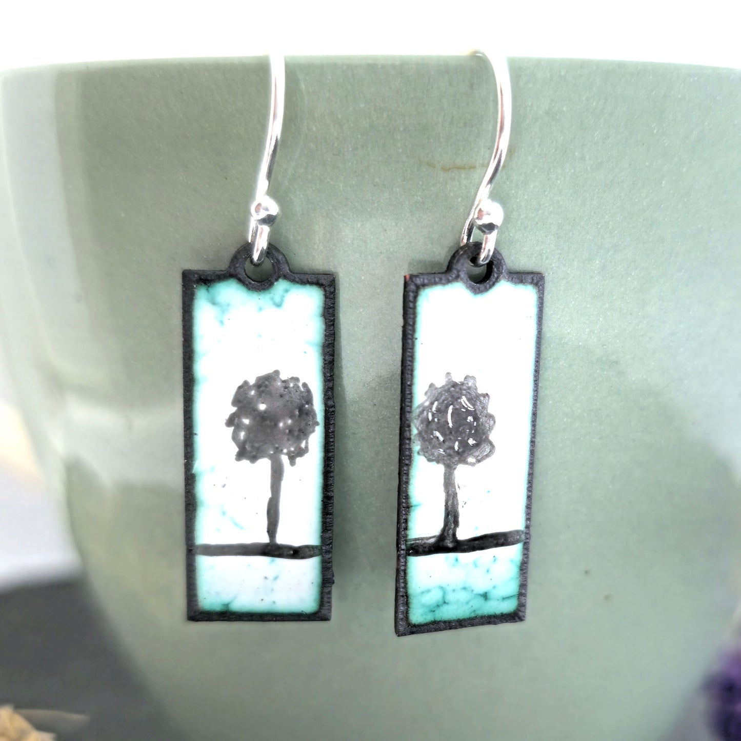 Rectangle enamel earrings with a single tree silhouette on each earring, in white, green and black, suspended from silver hooks. On a pale green background. Handmade. Mijoux Jewellery.