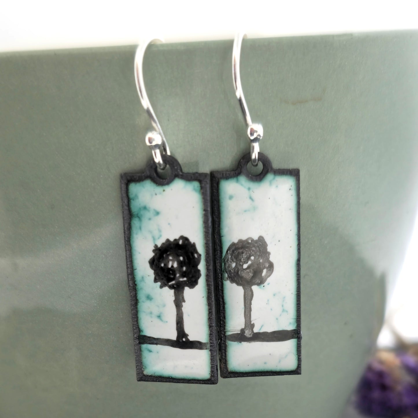 Rectangle enamel earrings with a single tree silhouette on each earring, in white, green and black, suspended from silver hooks. On a pale green background. Handmade. Mijoux Jewellery.
