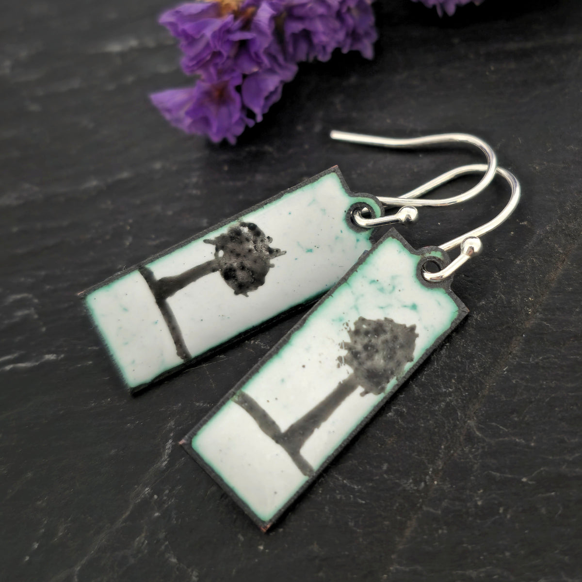 Rectangle enamel earrings with a single tree silhouette on each earring on a white background with green accents, suspended from silver hooks. On a black background with flowers. Handmade. Mijoux Jewellery.