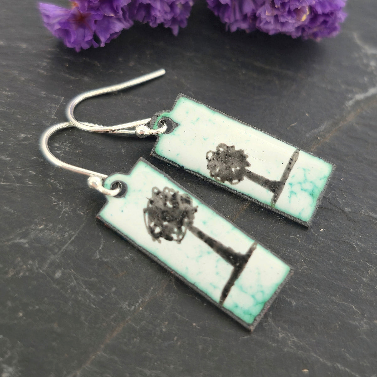 Rectangle enamel earrings with a single tree silhouette on each earring on a white background with green accents, suspended from silver hooks. On a black background with flowers. Handmade. Mijoux Jewellery.