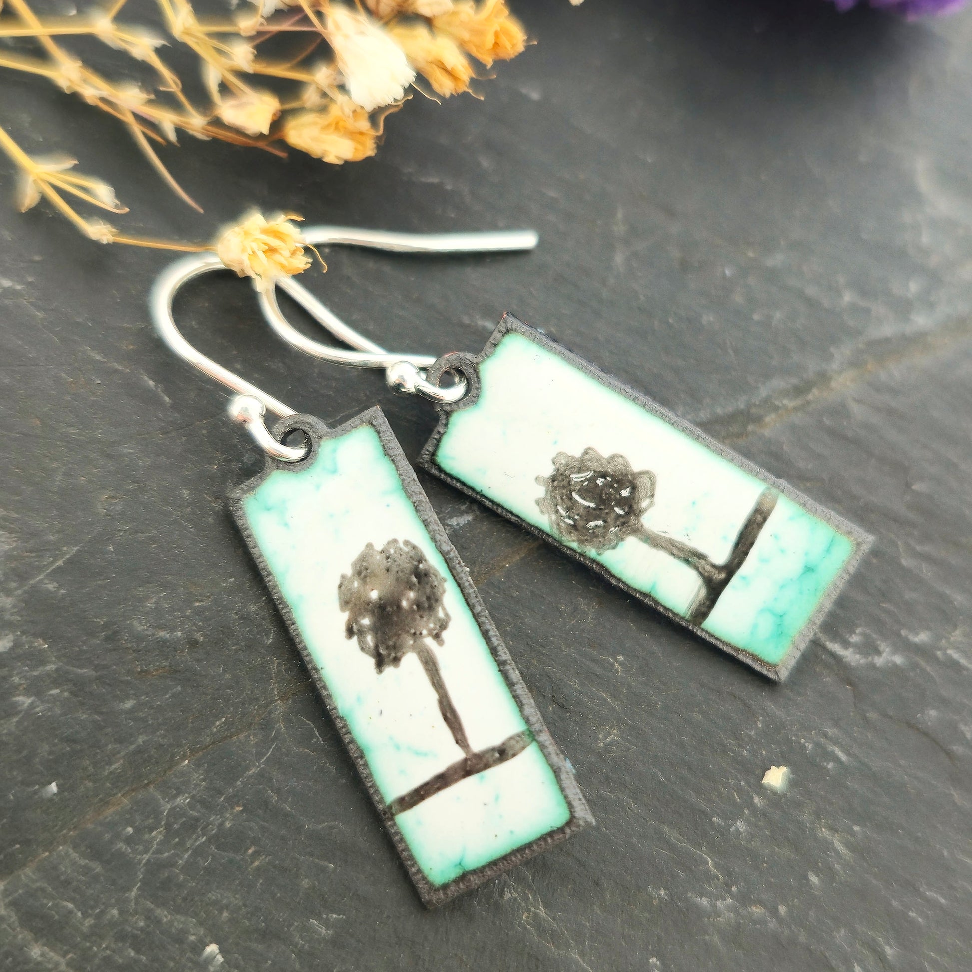 Rectangle enamel earrings with a single tree silhouette on each earring suspended from silver hooks.On a black background with flowers. Handmade. Mijoux Jewellery.