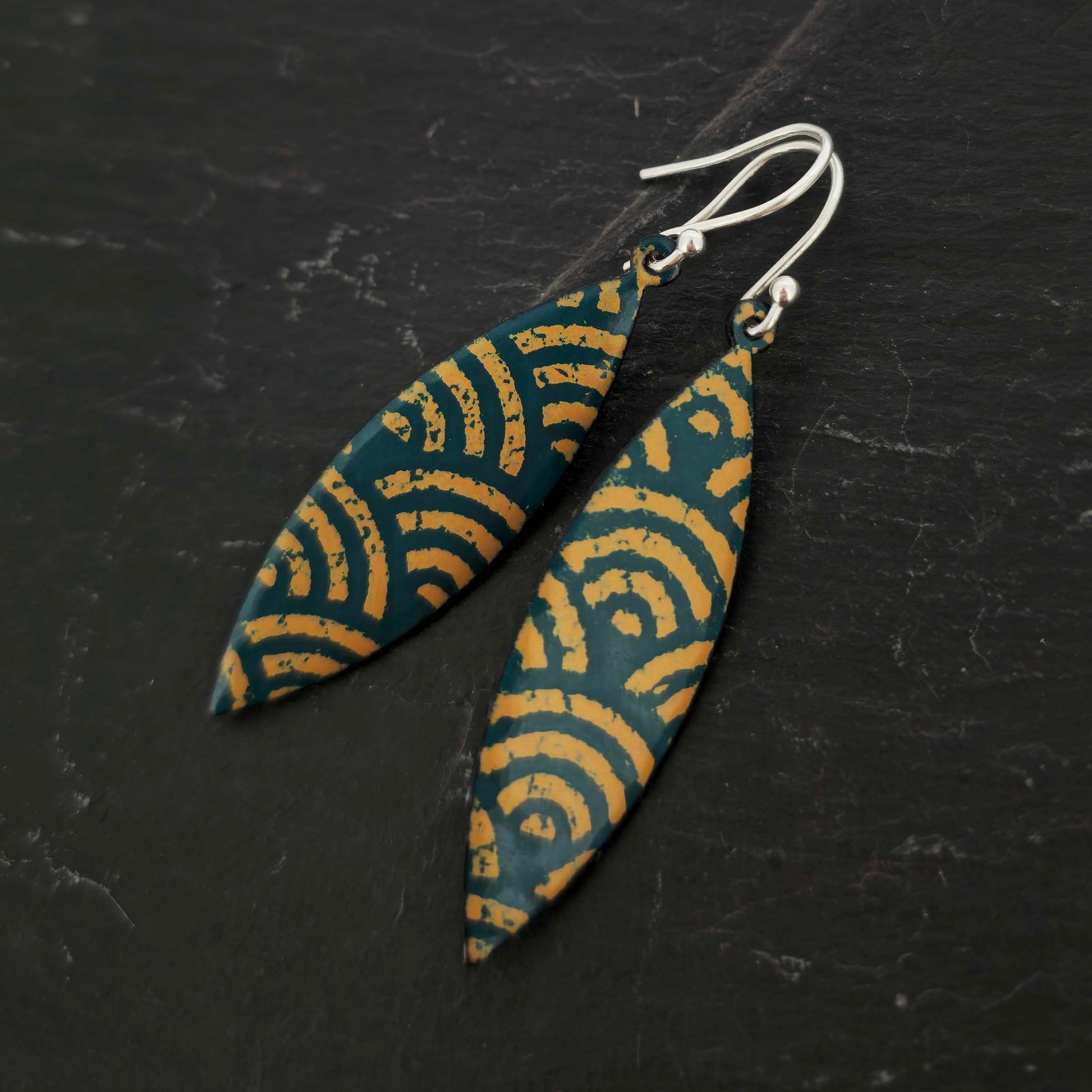 A pair of boat shaped drop earrings with a dark green background and an art deco style fan pattern in gold enamel. On silver ear hooks. Shown on slate.
