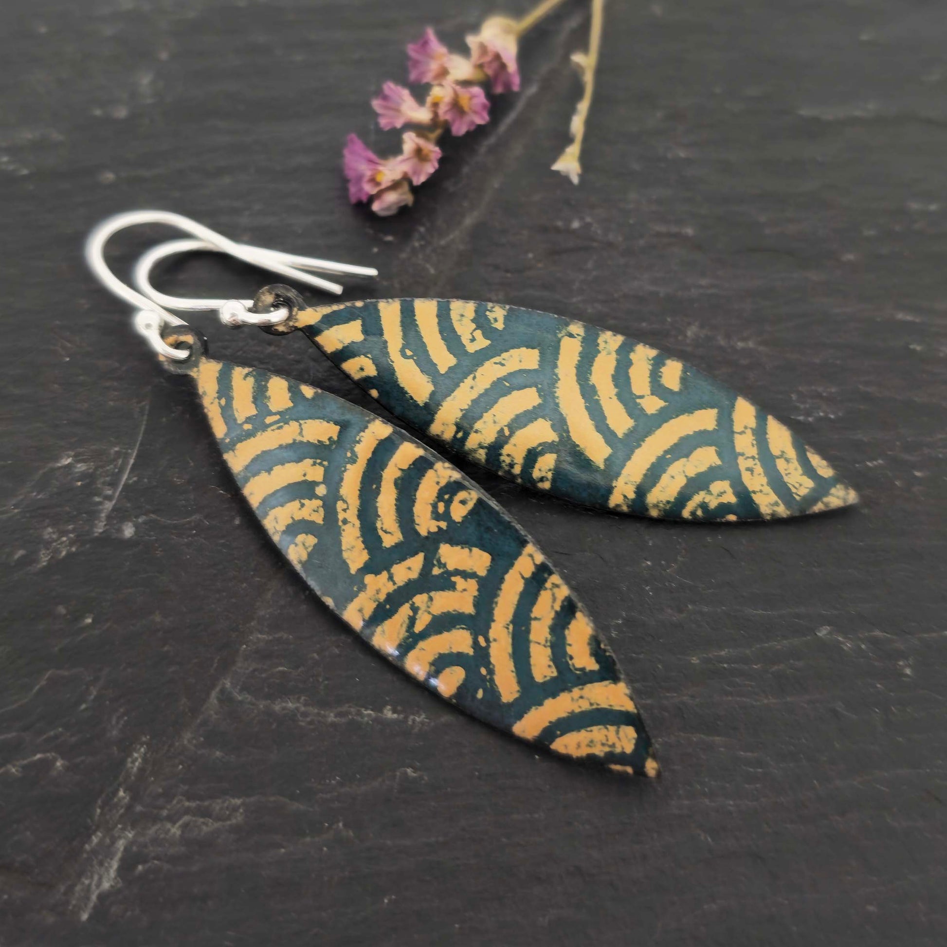 A pair of boat shaped drop earrings with a dark green background and an art deco style fan pattern in gold enamel. On silver ear hooks. Shown on slate with flowers.