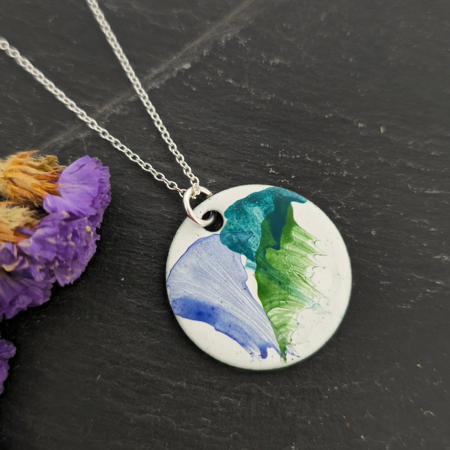 A round enamel pendant with a white background and splashes of dark blue, dark green and jade green. Suspended from a silver chain. On a black background with purple flowers.Mijoux Creations.