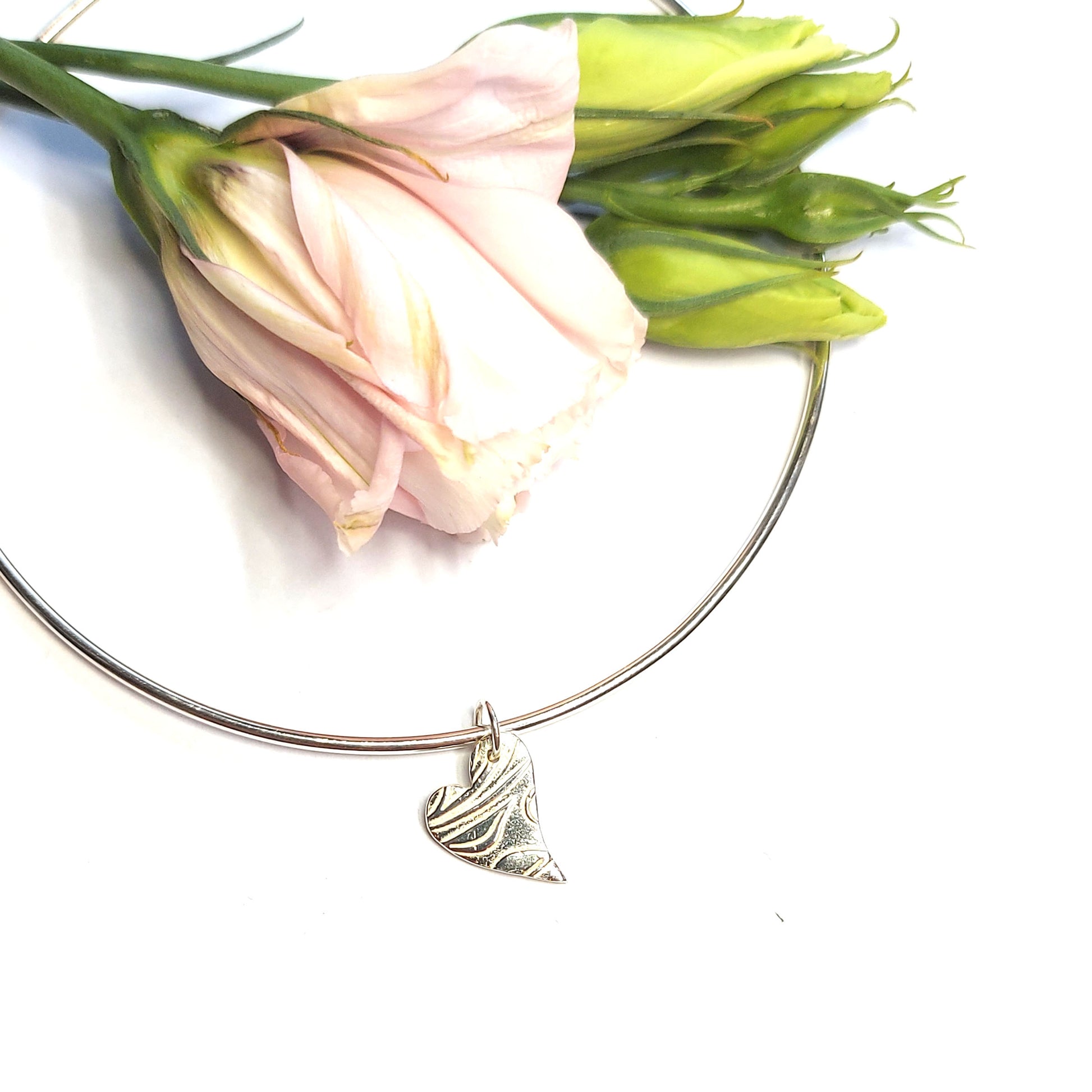A round silver bangle with an asymmetrical patterned heart charm. Pictured with flowers
