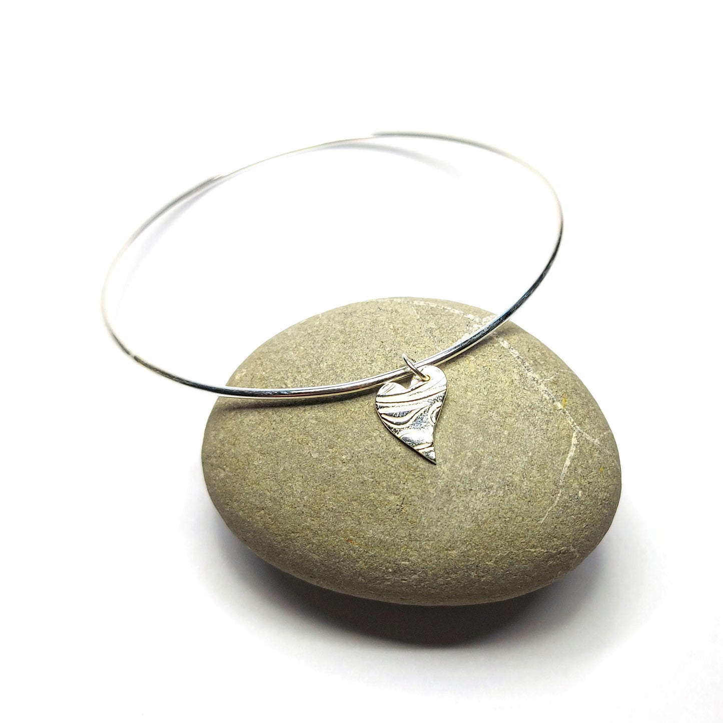 A round silver bangle with an asymmetrical patterned heart charm. Pictured on a pebble.