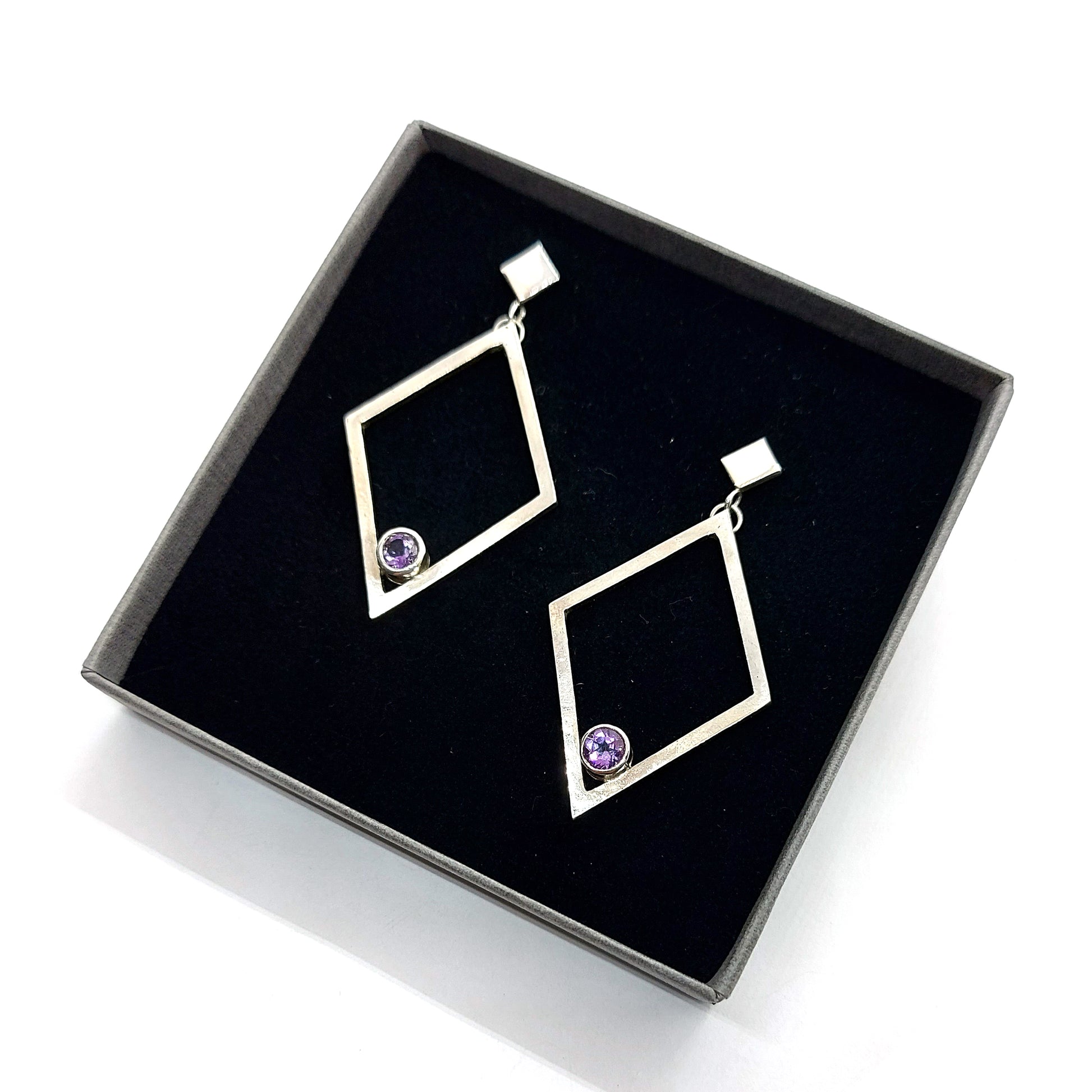 A pair of silver statement earrings featuring a top diamond shaped stud with a large open diamond shape drop beneath. Inside at the bottom of the large drop diamond is a round purple amethyst. Pictured in a box.